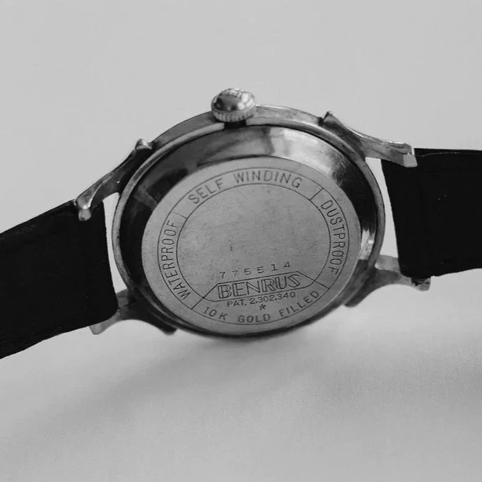 Back side of a vintage Benrus wristwatch with a metal case and black strap, showing engraved model and patent information.