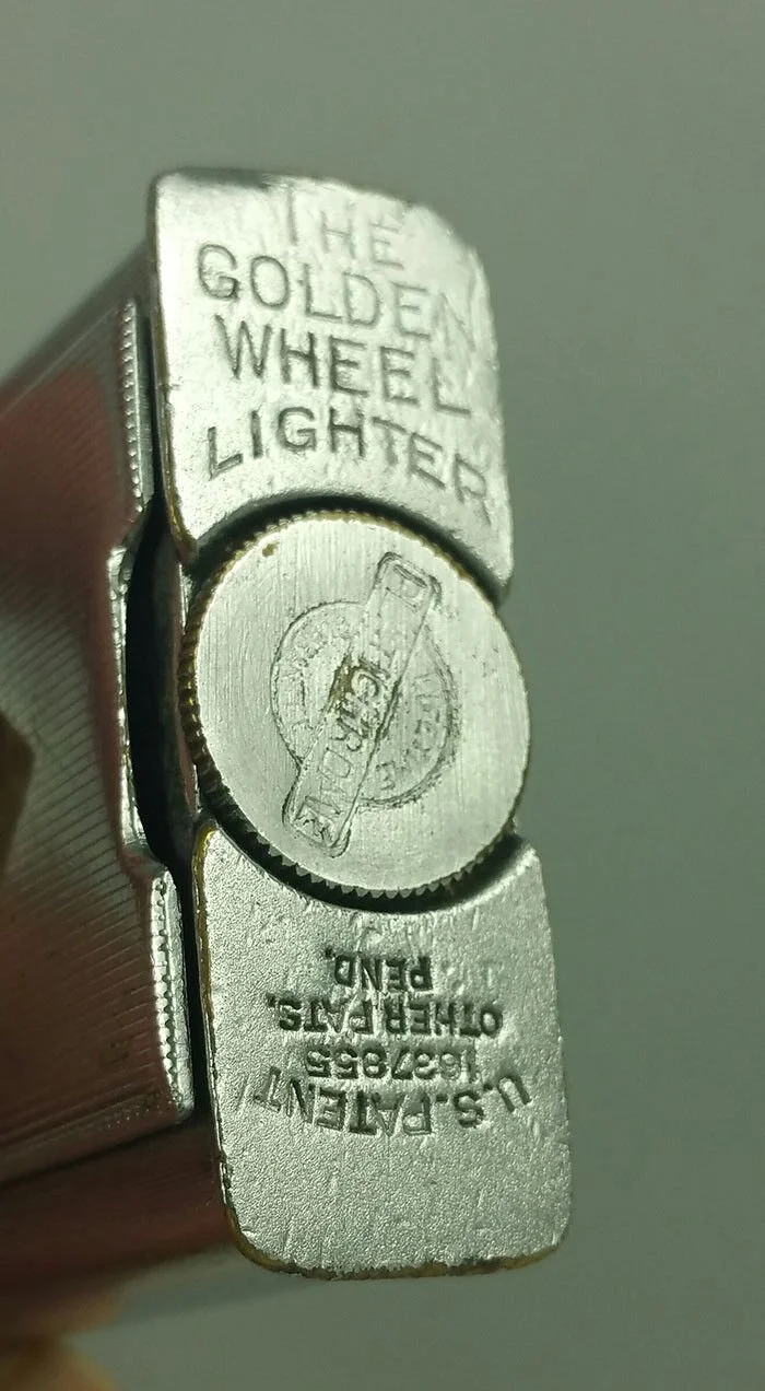 Close-up of a silver Zippo lighter with engraved text 'The Golden Wheel Lighter' and a circular logo with a dollar bill illustration, and additional text 'U.S. Patent 1627/9555 Other Patents Pending'.
