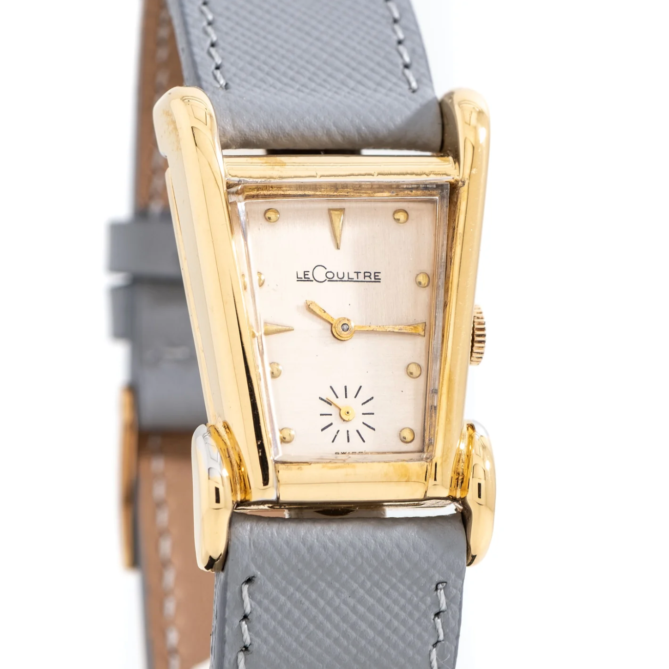 A vintage LeCoultre wristwatch with a rectangular gold case, silver dial, gold hands, and a small seconds subdial at 6 o'clock, attached to a grey leather strap.