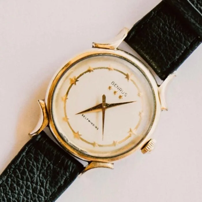 A vintage-style wristwatch with a black leather band and a round, gold-toned case. The watch face is white with gold stars and a decorative border. The watch hands are black, with the hour and minute hands pointing to approximately 9:15.