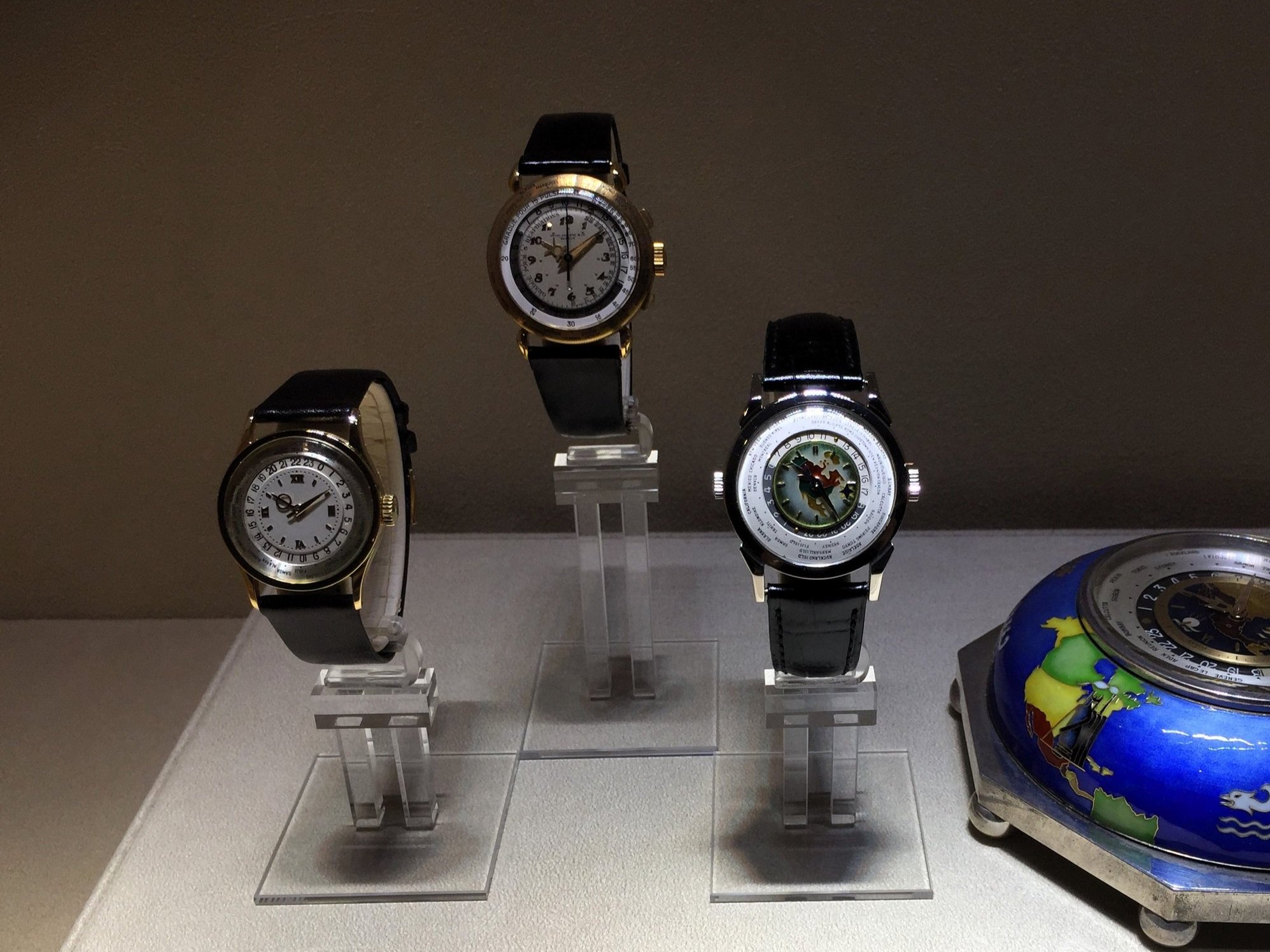 Display of four luxury watches on clear stands, with a globe-shaped object partially visible on the right.