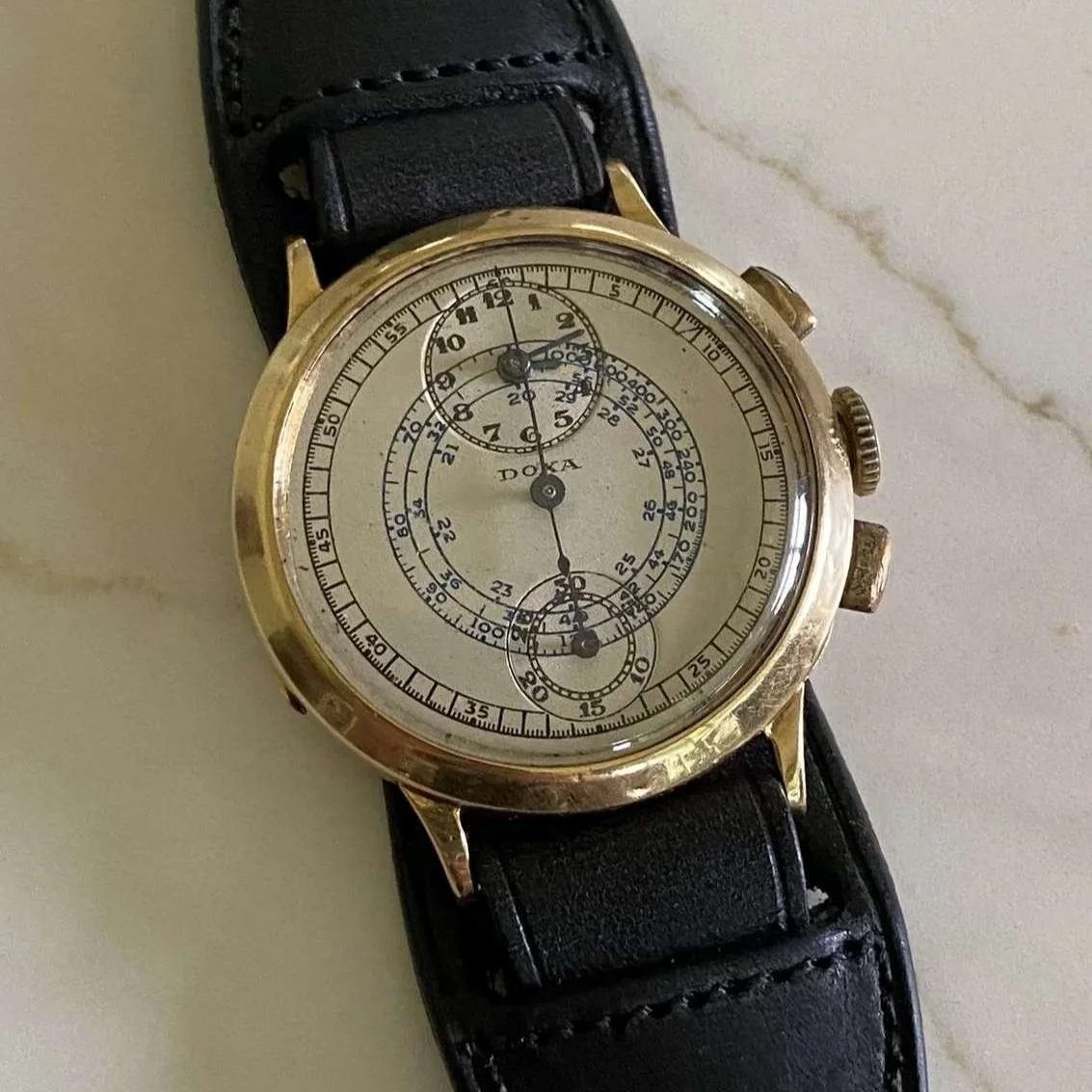 A wristwatch with a gold case and black leather strap, featuring a detailed chronograph dial with multiple subdials and markings.