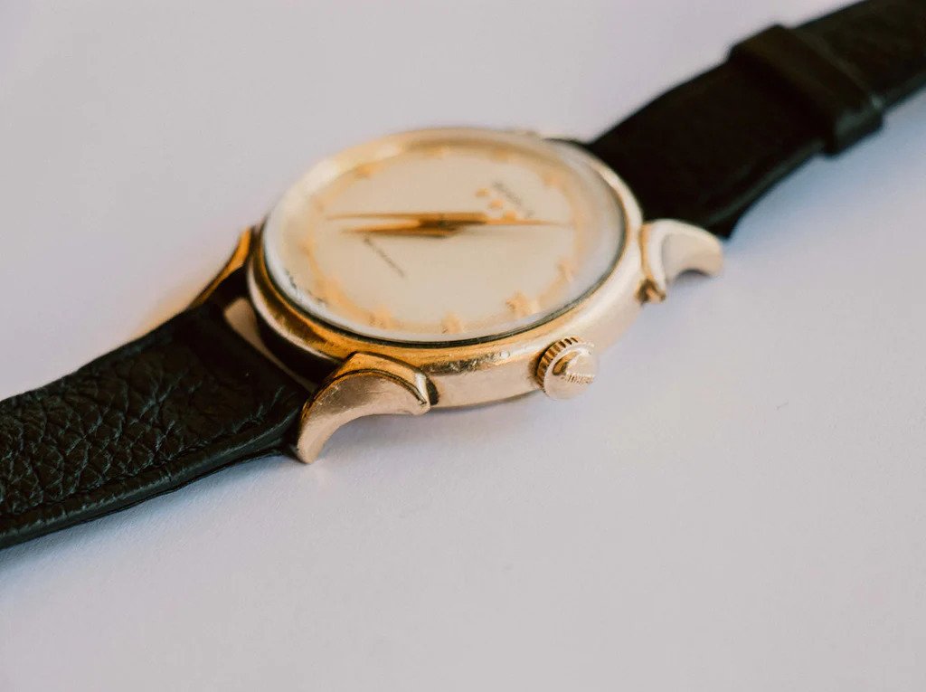 A vintage wristwatch with a round gold-colored case, a white dial, and a black leather strap, lying on a white surface.