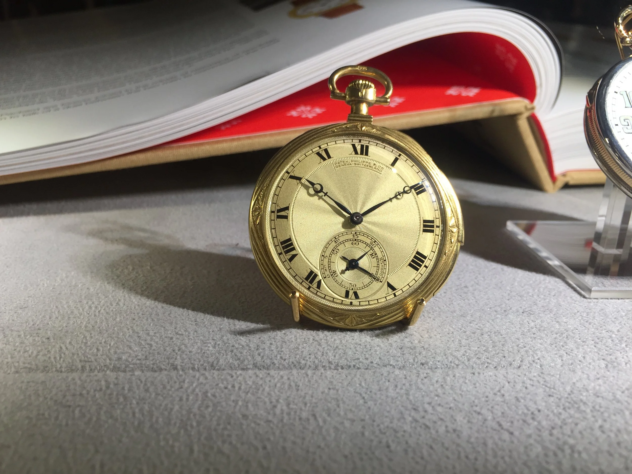 Close-up of a gold pocket watch with Roman numerals, placed on a gray surface in front of an open book with a red and white page edge, and another watch partially visible.