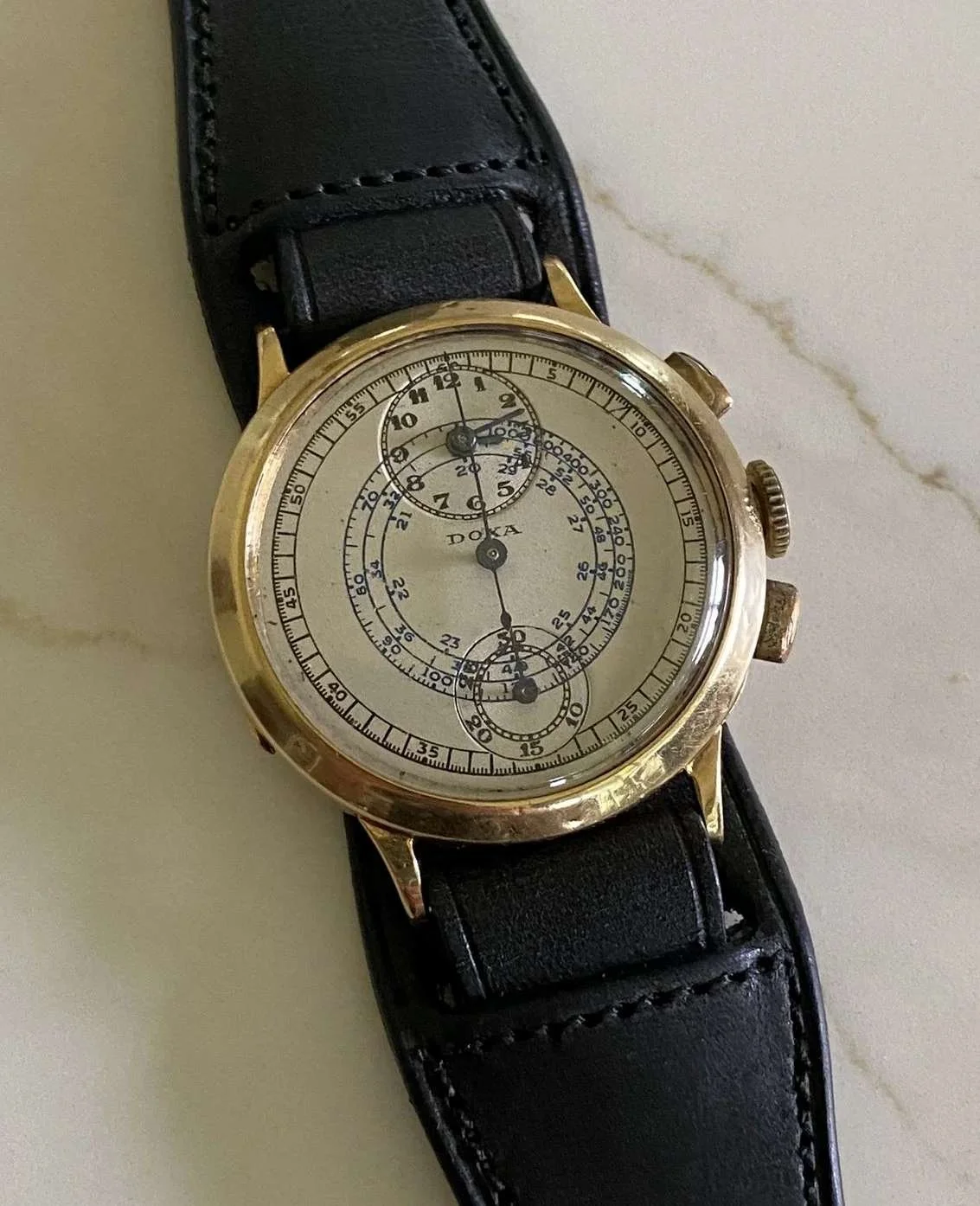 A vintage wristwatch with a black leather strap, gold case, and a detailed chronograph face displaying multiple dials and markers.