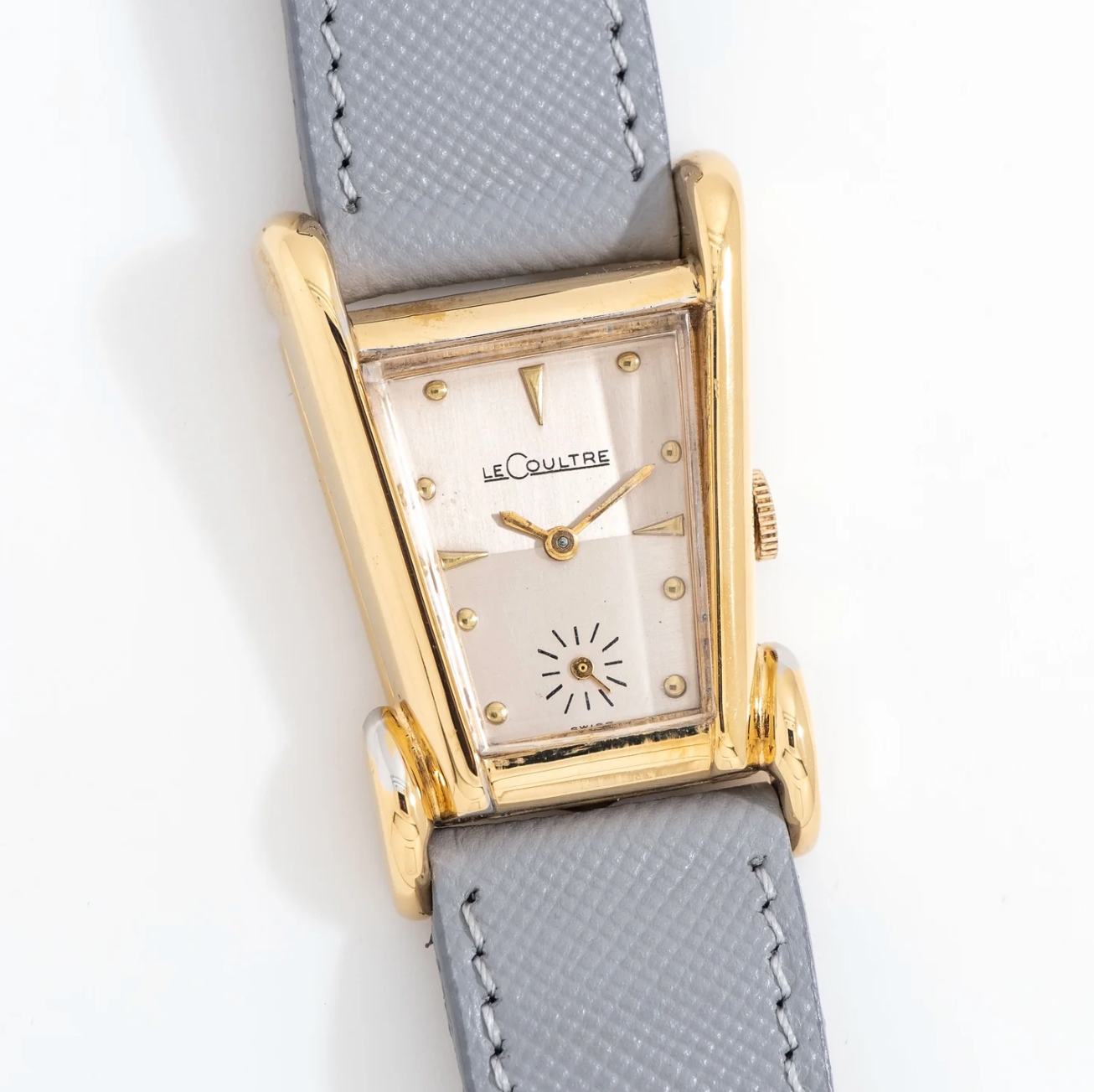 A vintage LeCoultre wristwatch with a rectangular gold case, a white dial, gold hour markers, and gray leather strap.