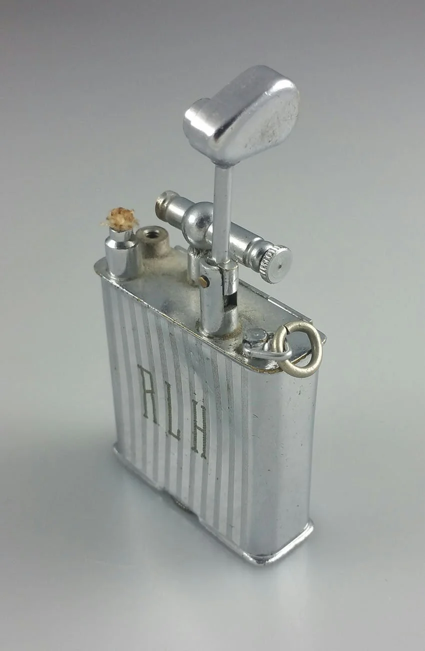 Close-up of a shiny silver zippo lighter with a small piece of tobacco or debris on the side, set against a plain background.