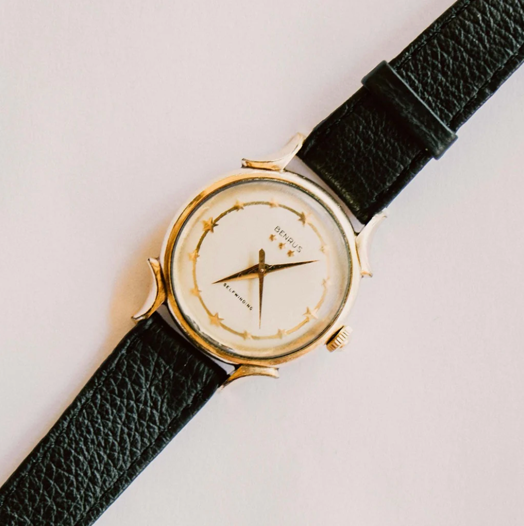 A vintage wristwatch with a gold case, black leather strap, and an off-white dial featuring star and moon designs, with the brand name "BENUES" on the face.