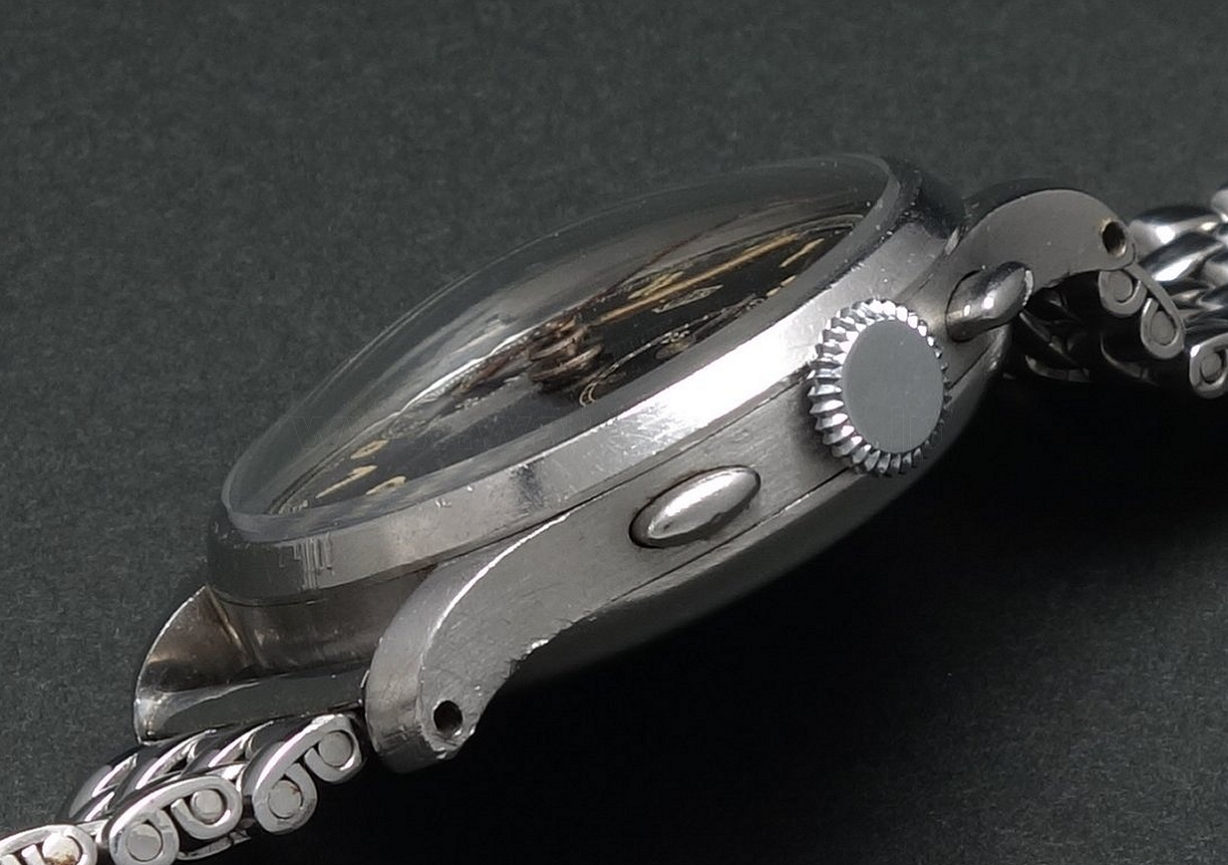 Close-up of a vintage stainless steel wristwatch with a metal link band, featuring round and oval buttons and a textured crown on a black background.