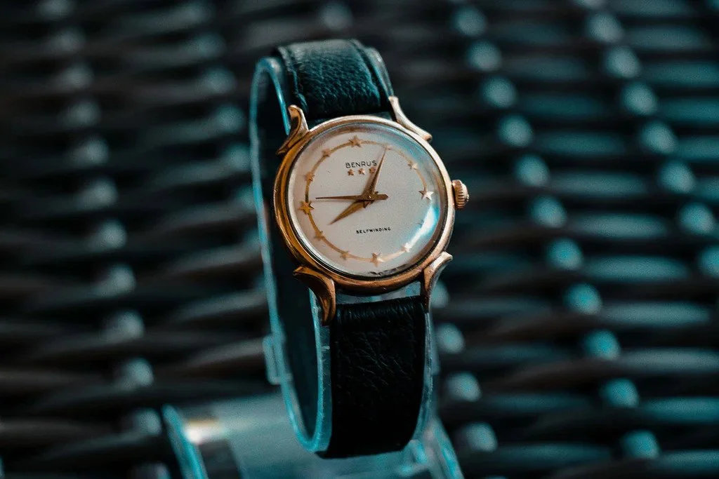 A wristwatch with a black leather strap and a round gold case, displaying a white dial with gold star hour markers and gold hands, placed against a dark textured background.