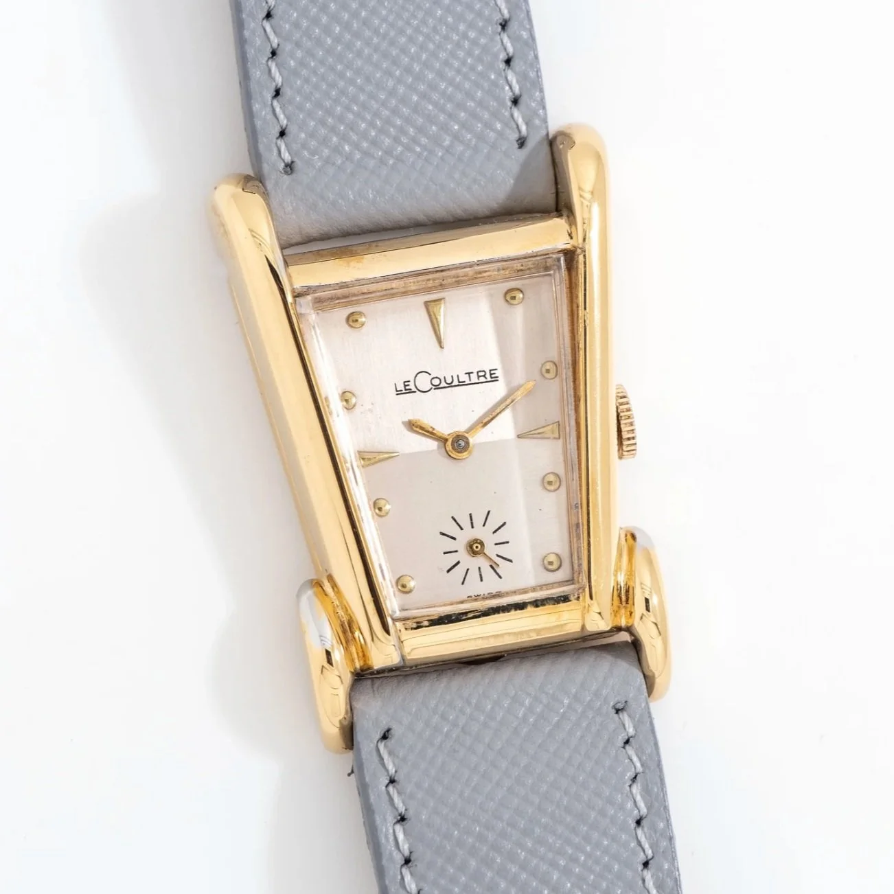 A vintage LeCoultre wristwatch with a rectangular gold case, white dial, gold hands, and a small seconds subdial at the 6 o'clock position, paired with a light gray leather strap.