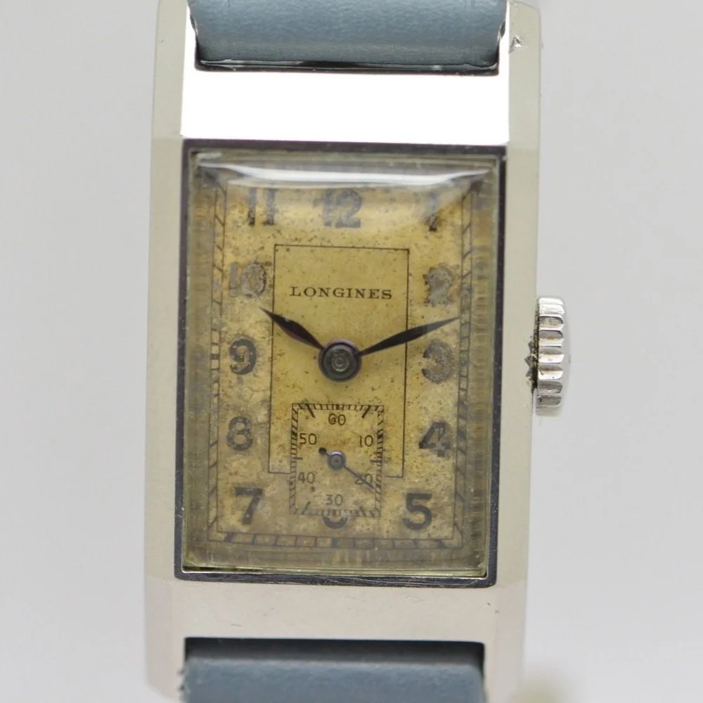 Vintage Longines wristwatch with a rectangular metal case, yellowed aged dial, black numerals, and black hour and minute hands. The dial includes a small seconds subdial at 6 o'clock and an outer minute track. The watch has a crown on the right side and is attached to a gray strap.