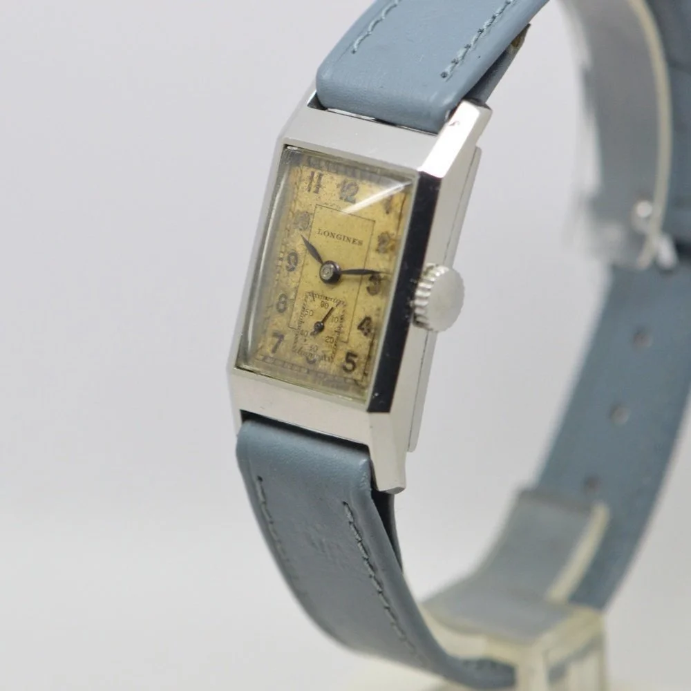 Vintage Longines wristwatch with a rectangular face, yellowed dial, black numbers, small seconds subdial, and a light blue leather strap.