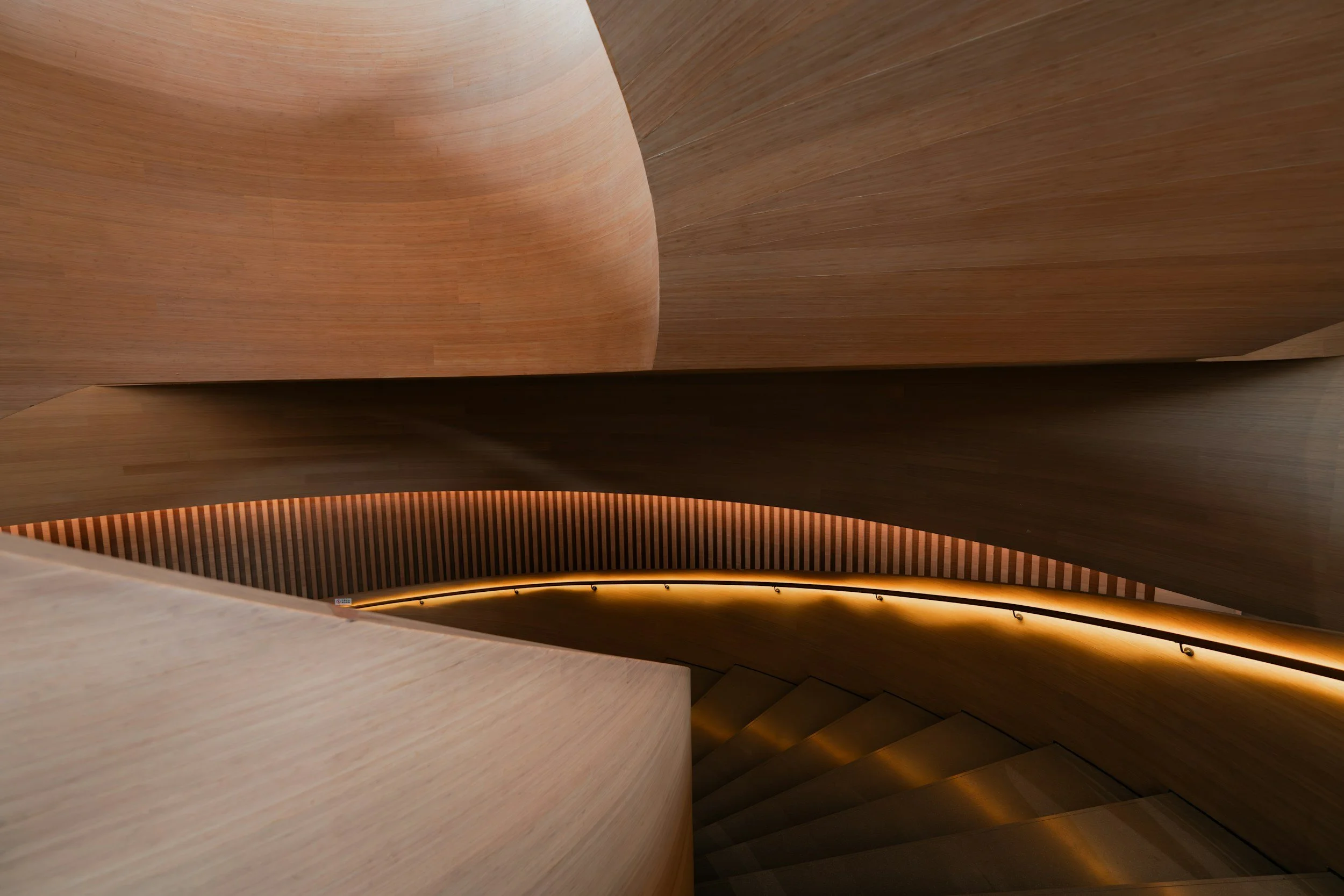 Interior view of a modern architectural staircase with curved wooden walls and railings, featuring warm lighting.