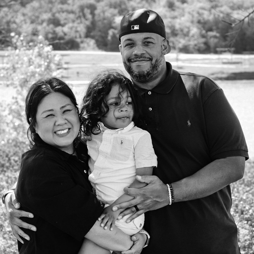 A family of three outdoors, smiling and hugging, with a field and trees in the background.