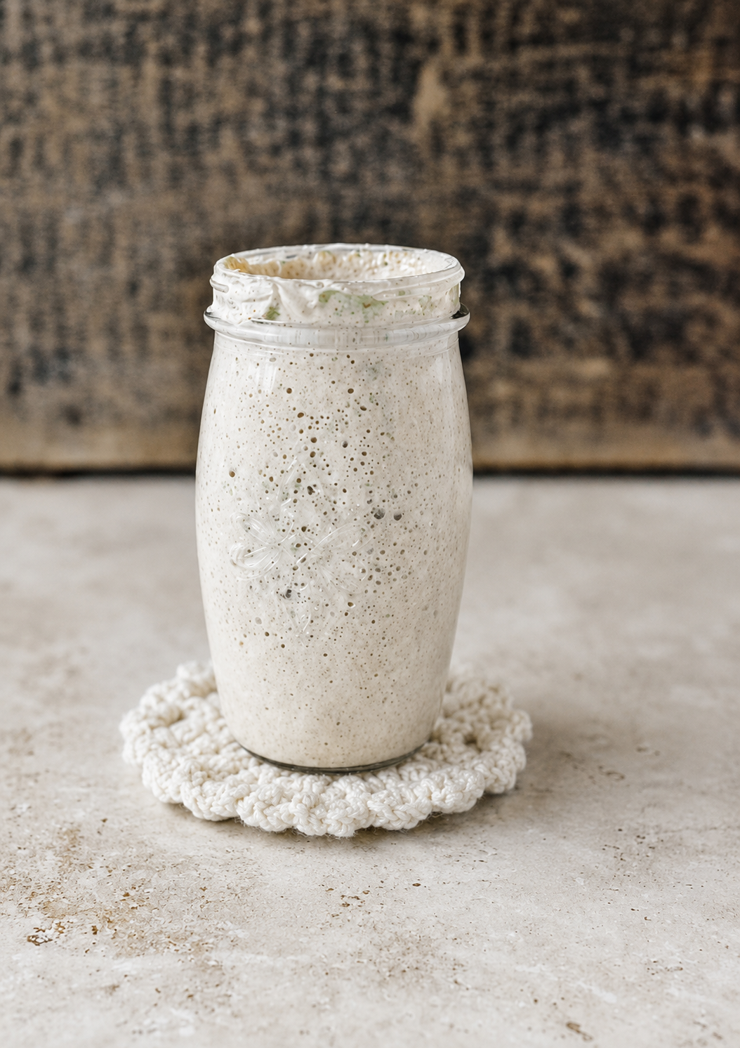 A glass jar filled with an off-white, frothy smoothie with visible green flecks, placed on a crocheted coaster on a light-colored surface with a rustic, wooden background.