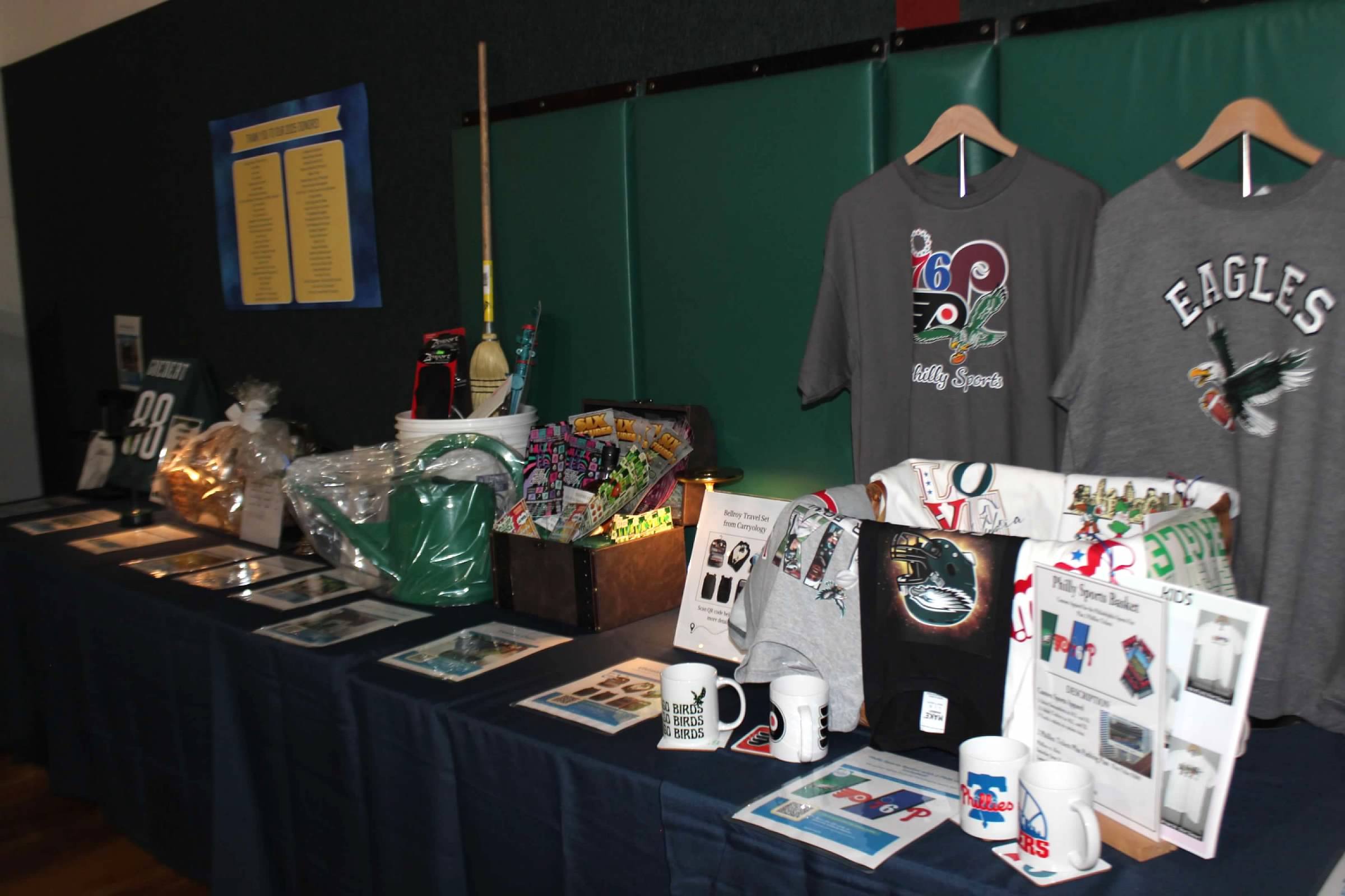 A display table with Philadelphia Eagles-themed merchandise, including T-shirts, mugs, and other items, in an indoor setting with a dark wall background and a green panel.