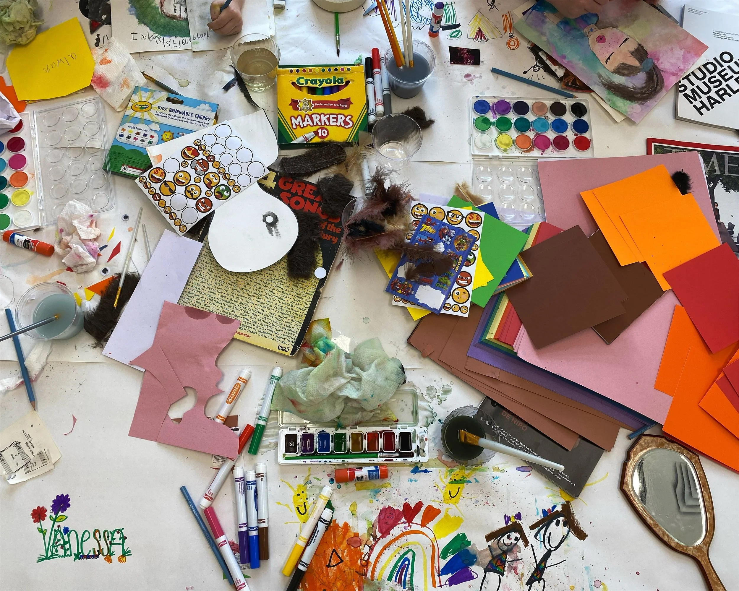 A cluttered art table with various supplies including paper, markers, paint, brushes, stickers, and colored paper sheets. There are small artworks, including a painted face and a rainbow drawing. The table surface is messy with multiple items scattered.