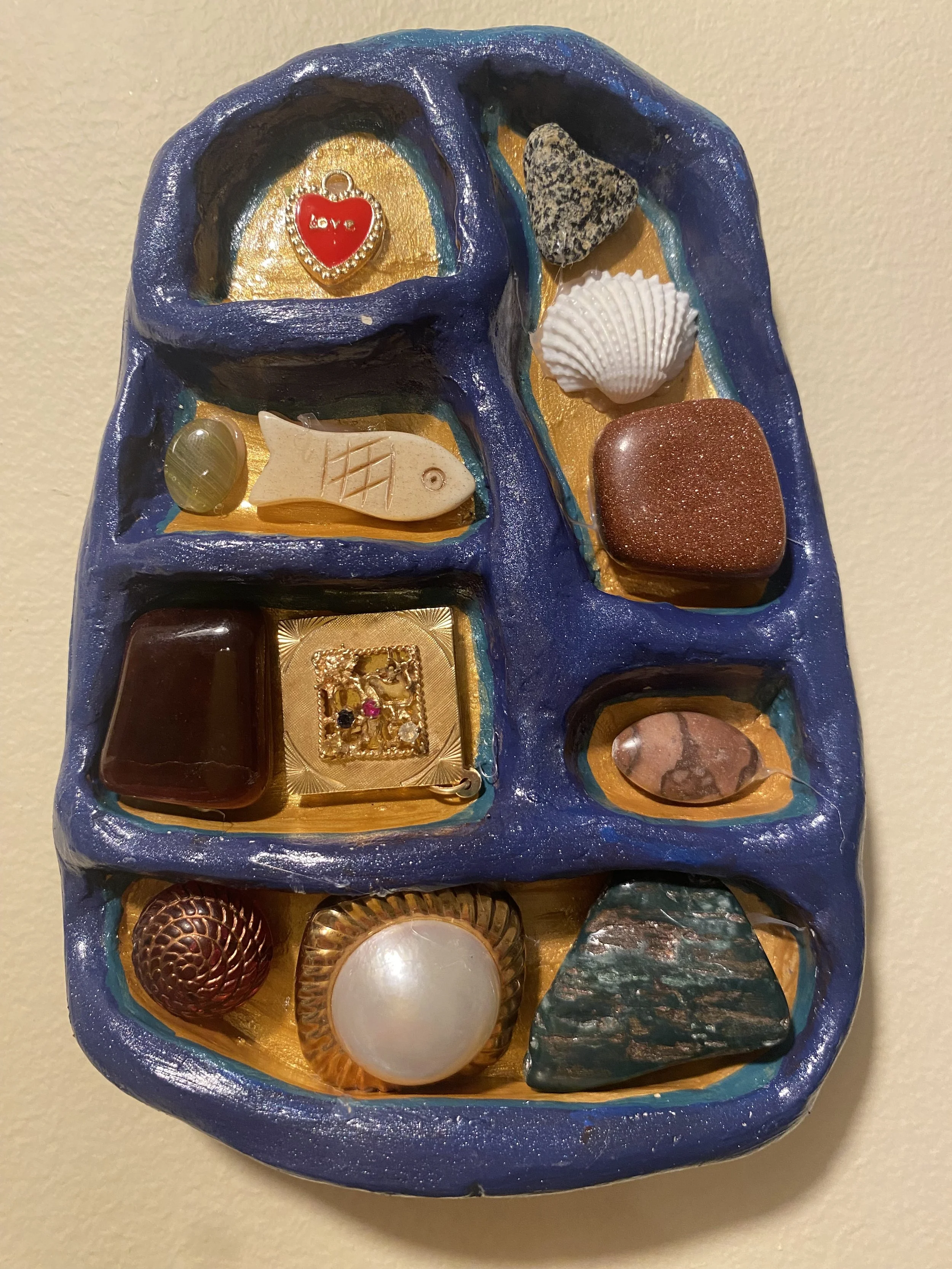 Decorative blue jewelry box with golden interior holding various jewelry items including a red heart-shaped pendant with the word 'love', a seashell, a stone, and other jewelry pieces.
