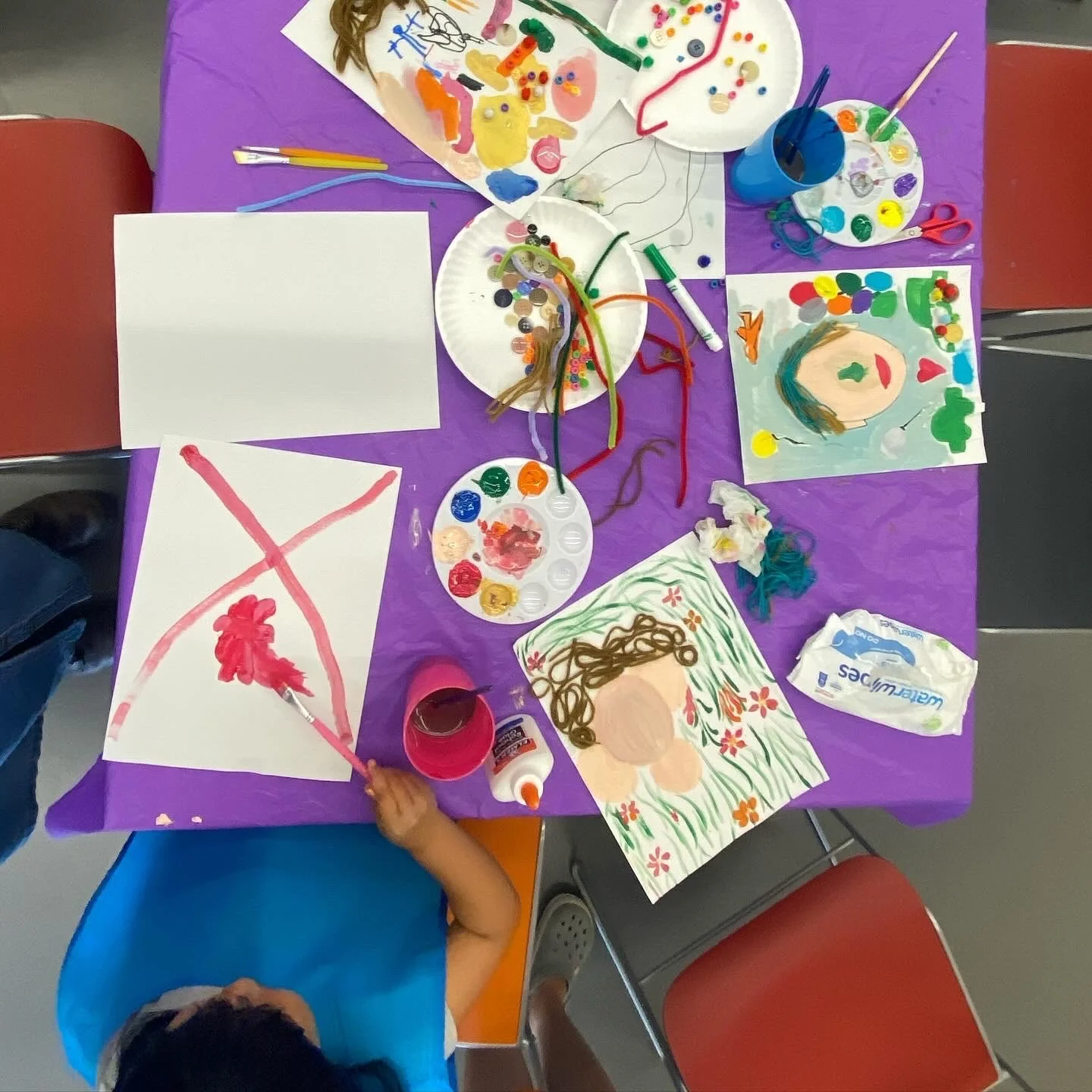 Overhead view of a child's art station with colorful paintings, paint palettes, brushes, and arts and crafts materials on a purple table covering. A child is painting on paper, with other completed artworks and supplies like glue and wet wipes on the table.