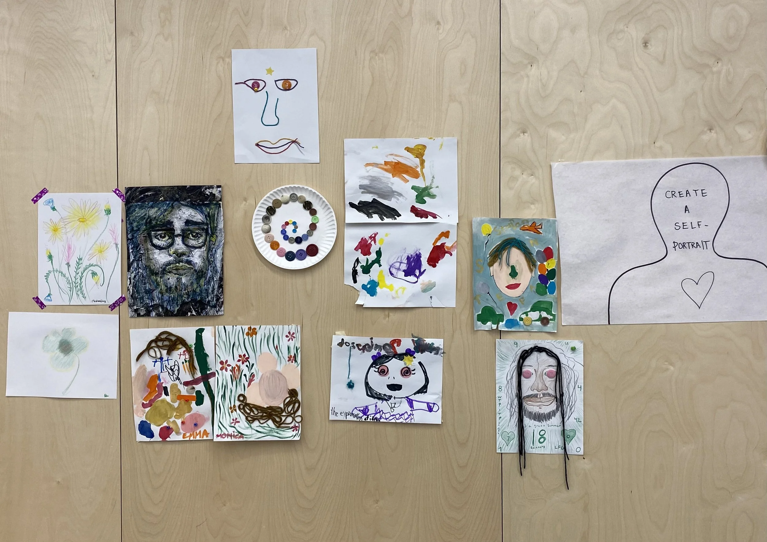 A wooden wall with various artistic drawings and paintings, including portraits, abstract art, a plate with colored buttons, and a silhouette with text "CREATE A SELF-PORTRAIT".