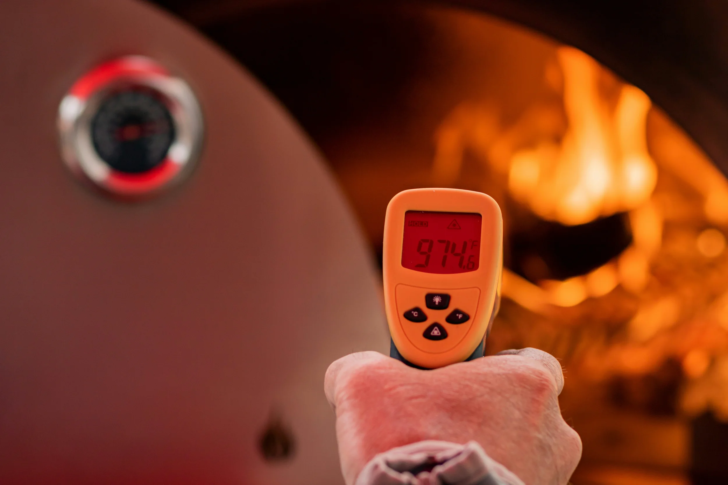 Close-up of a hand holding a digital infrared thermometer displaying a temperature of 94.7°F, with a blurred fireplace in the background.