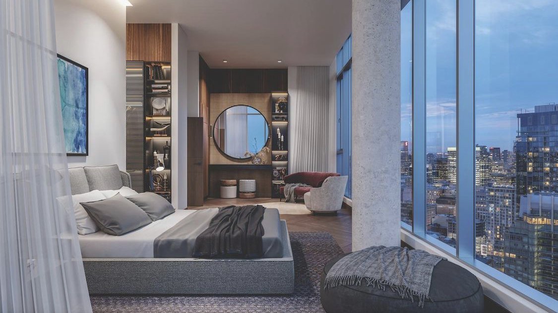 Modern bedroom with large windows showing a cityscape at dusk. Features a bed with gray bedding, an abstract wall art, a circular mirror, and a seating area with armchairs and a sofa.