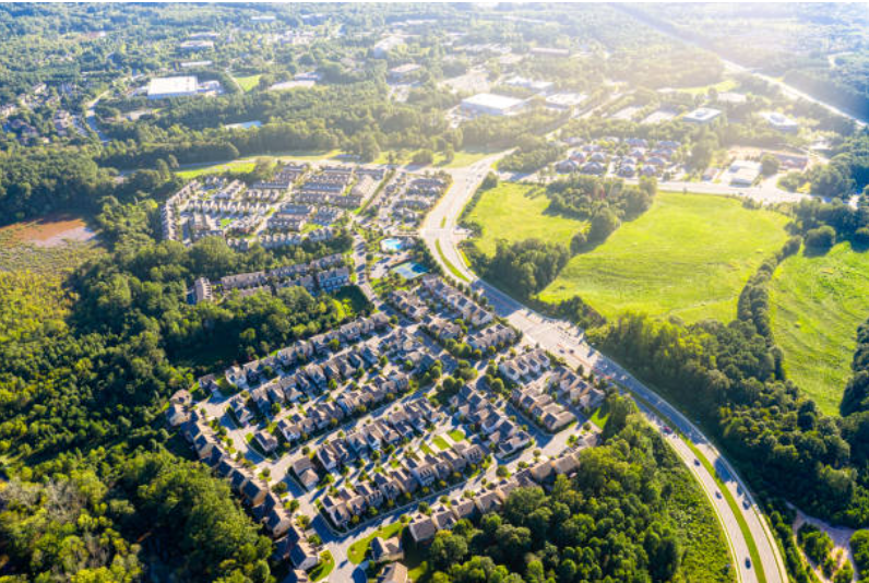 Aerial view of a suburban residential area with houses, roads, and green fields surrounded by trees.