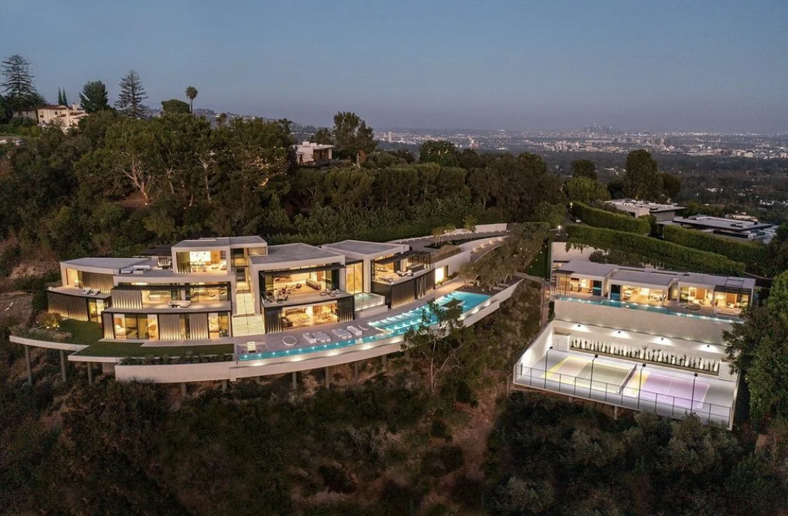 Luxury modern hillside home with illuminated outdoor pools, tennis court, and surrounding trees in Los Angeles.