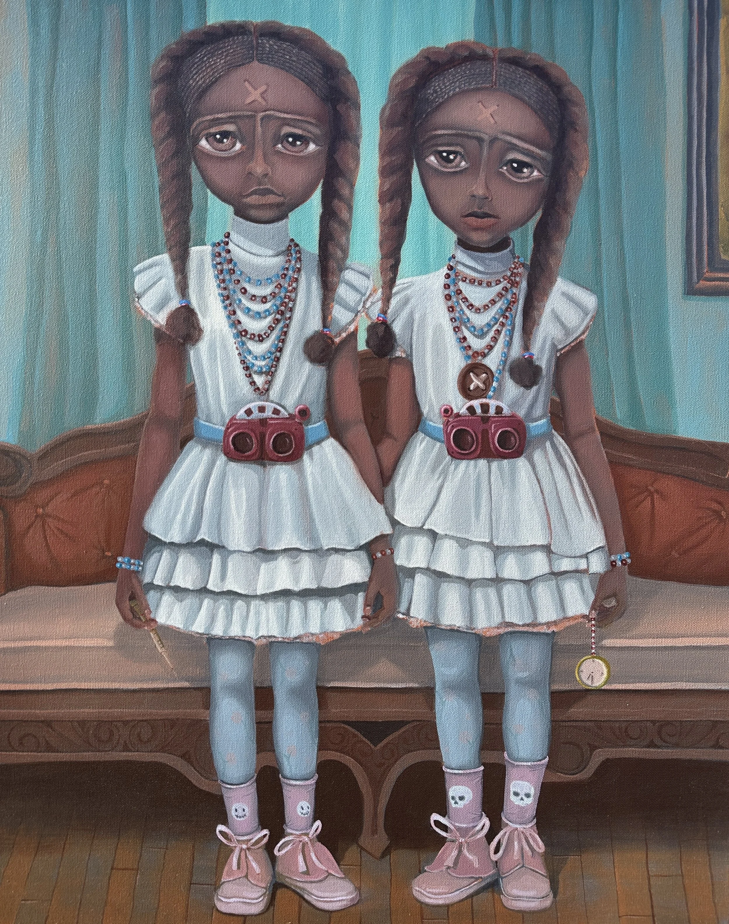 Two young girls with braided hair and dark skin, wearing white dresses, blue tights, and pink shoes with laces, standing in front of a wooden surface. One girl is holding a small stick with a yellow ring, and the other is holding a pink purse with a skull design. Both wear colorful beaded necklaces.