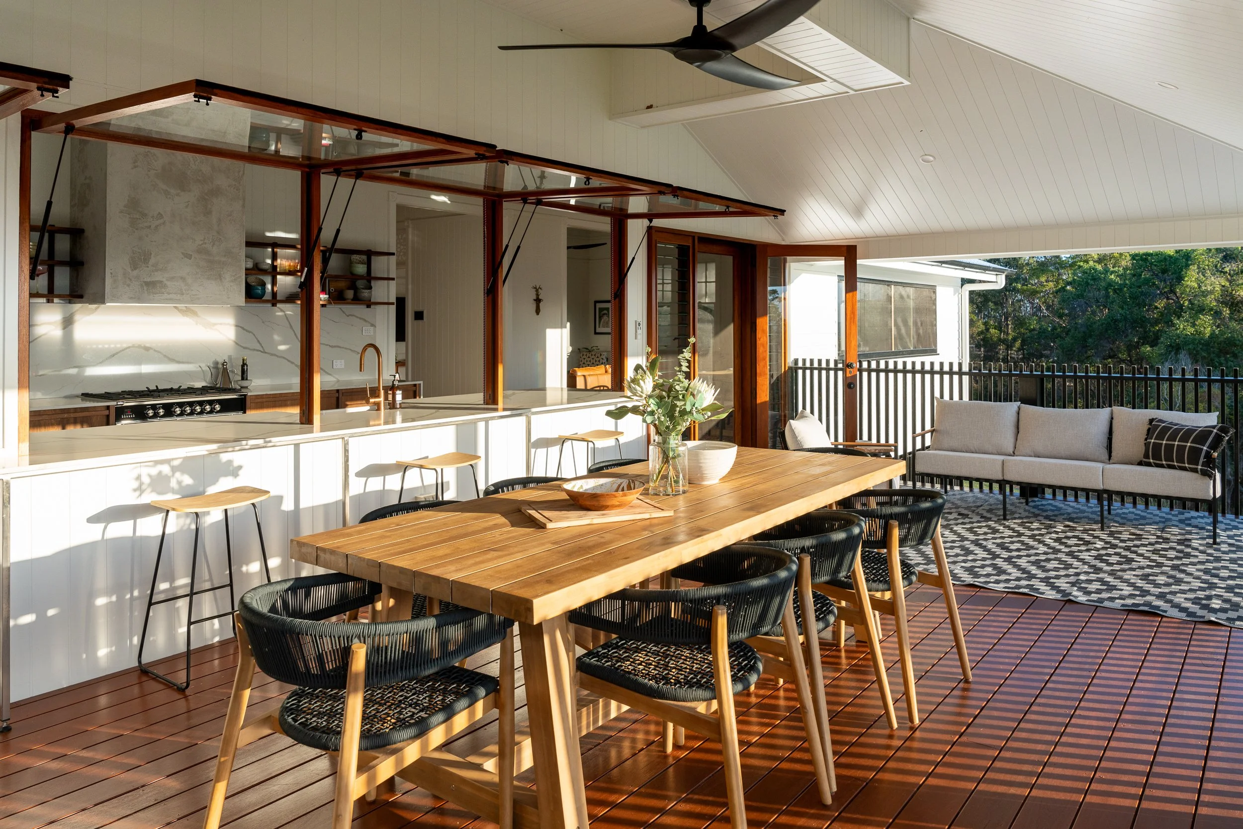 A spacious outdoor patio area with a large wooden dining table surrounded by black woven chairs, a cozy seating area with a sofa and cushions, and an open kitchen with a white marble island, open shelving with dishes, and a ceiling fan. The patio has