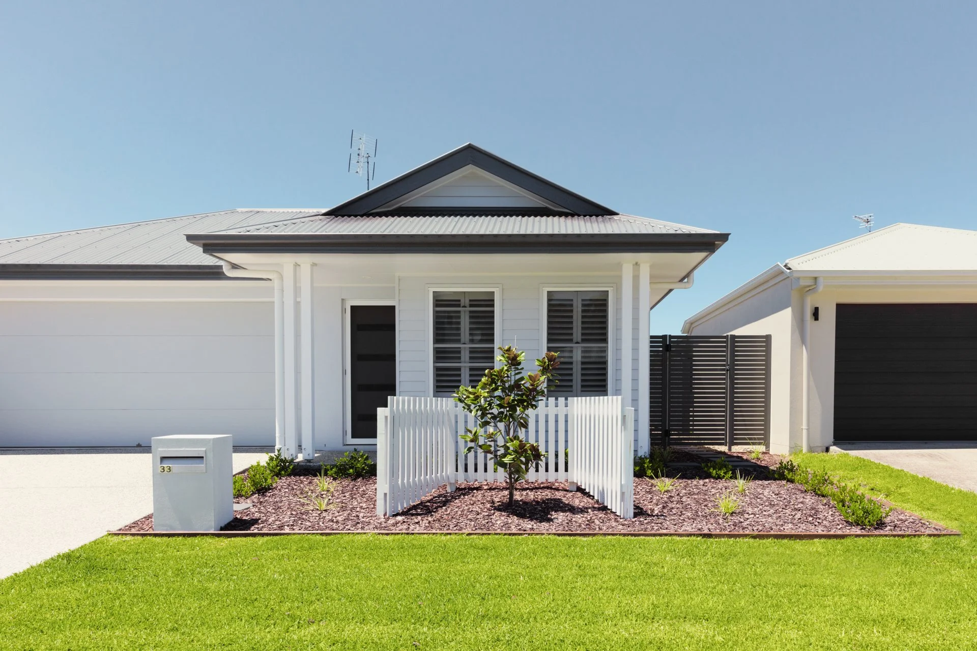 Front view of a modern white house with a gray roof, small garden, white picket fence, and a grassy lawn under a clear blue sky.