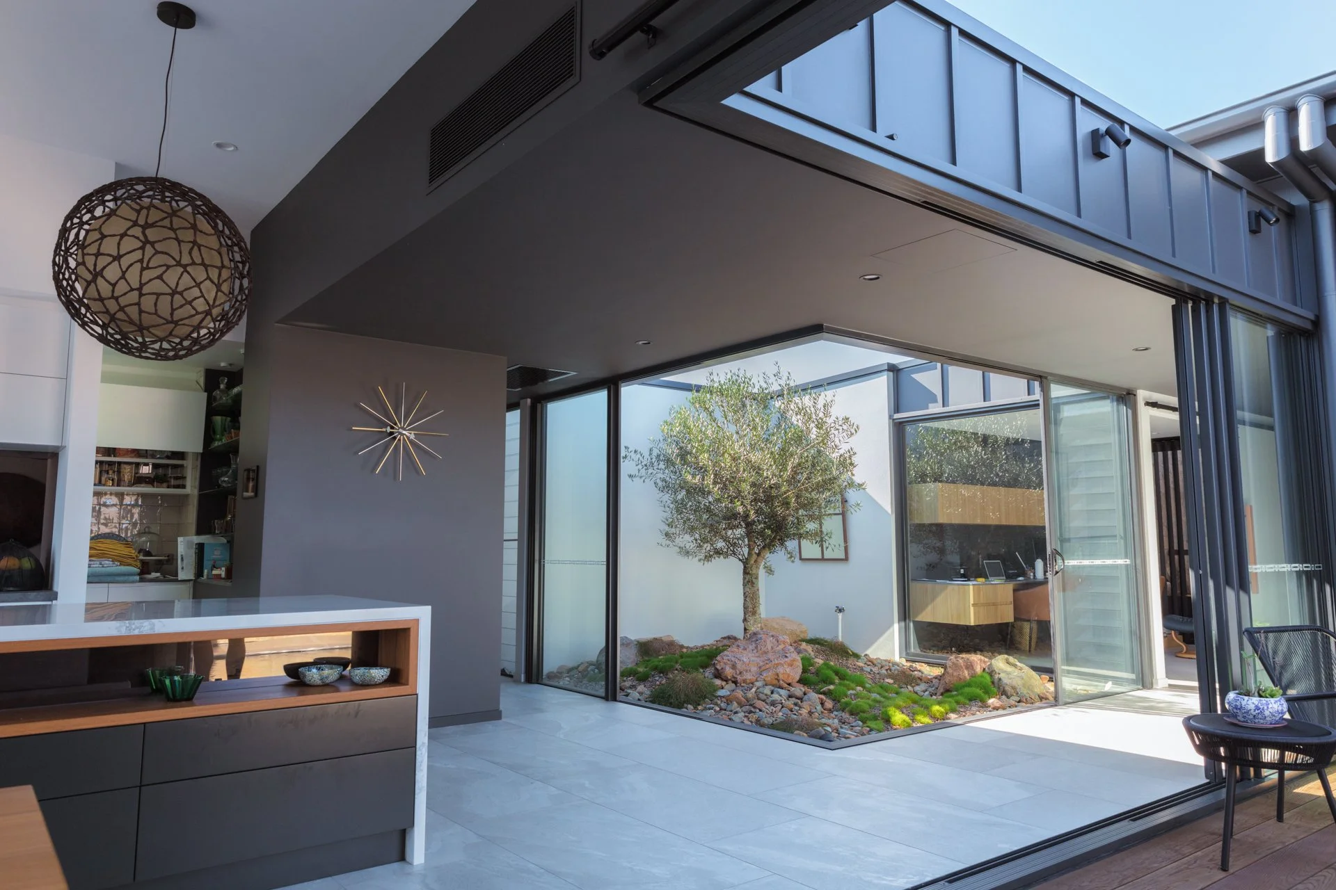 Modern interior living space with large glass sliding doors opening to a courtyard with a tree and rocks. Inside features a minimalistic design, including a hanging spherical light, a starburst wall clock, and built-in shelves.