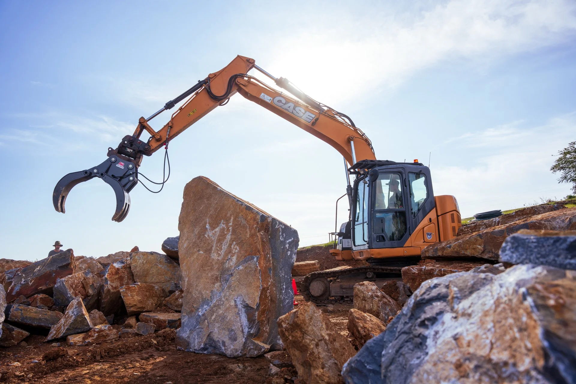 An orange excavator with the brand name 'CASE' on the arm, working outdoors on uneven terrain with large rocks. The sky is clear and blue, and the sun is shining brightly behind the excavator.