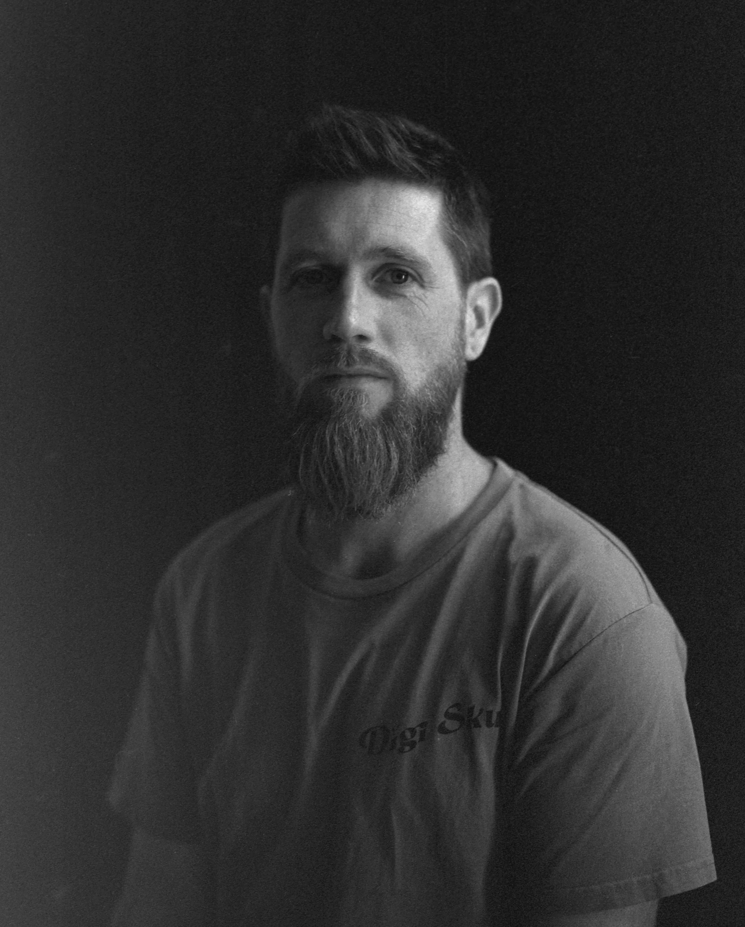 A black and white portrait of a man with a beard and styled short hair, wearing a T-shirt that says "Skin" and partially visible text.