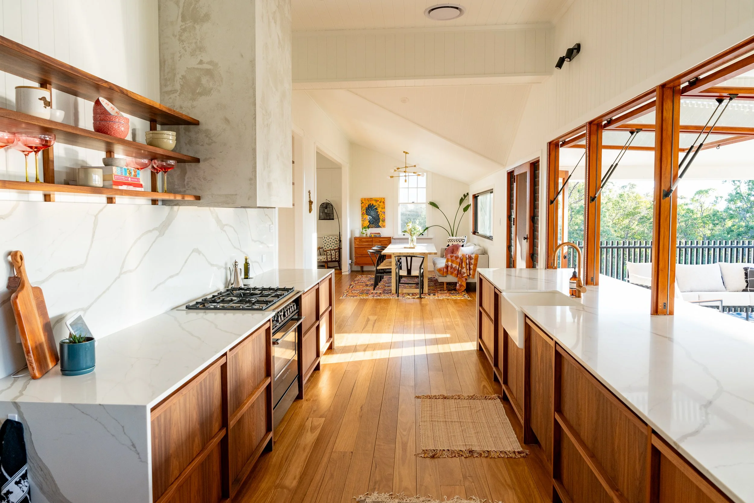 Open-concept kitchen and dining area with white quartz countertops, wooden cabinets, and large windows with a view of trees outside. The space features a breakfast bar, a gas stove, and a dining table with chairs, decorated with plants and artwork.