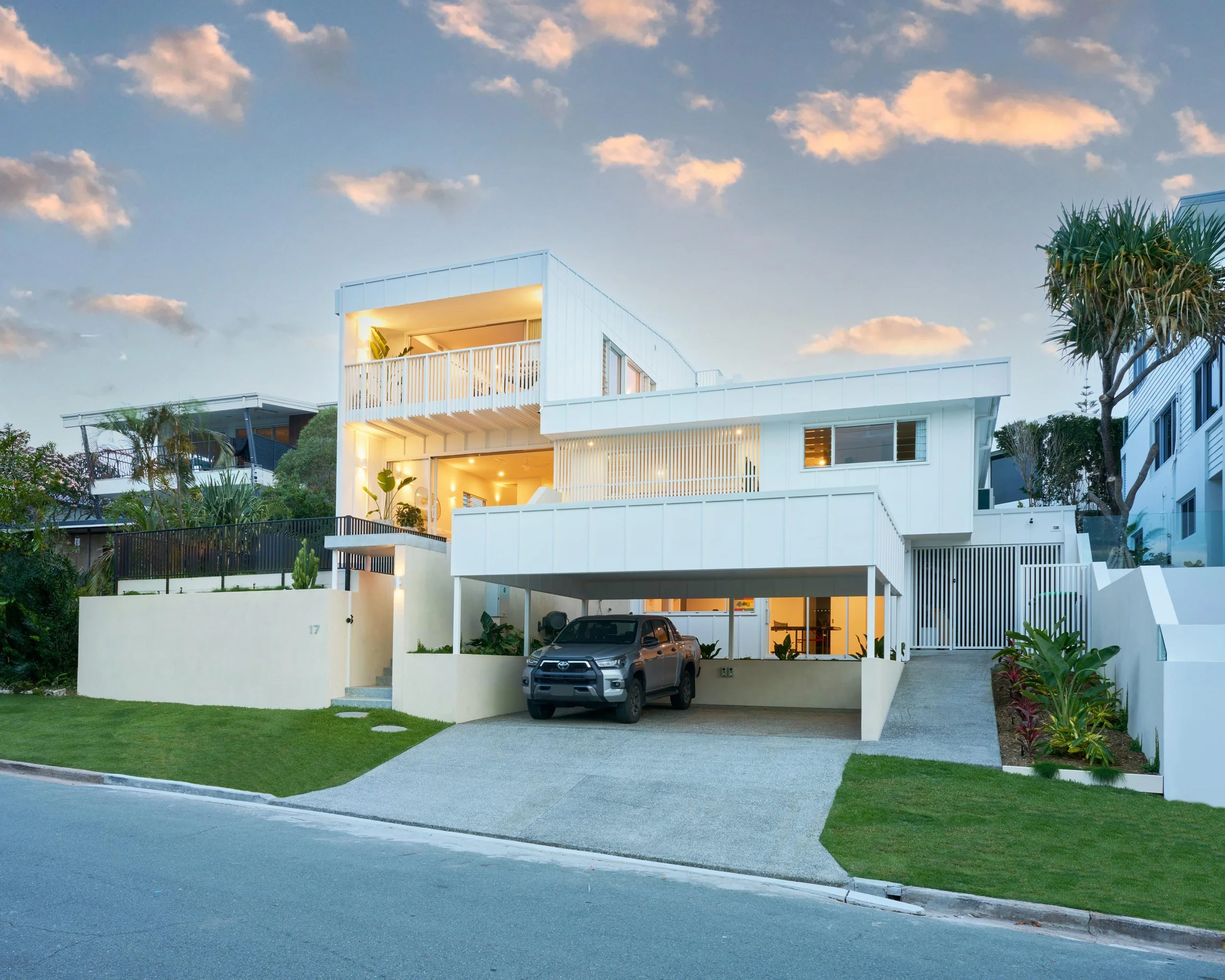 Modern white multi-story house with a driveway and a black SUV parked underneath a carport, surrounded by a green lawn and trees, with a cloudy sky in the background.