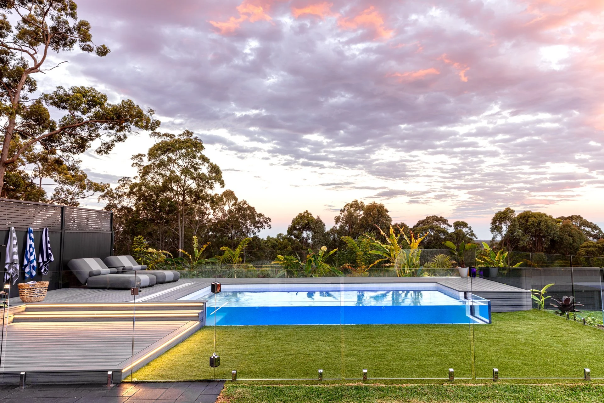 A backyard pool area at sunset with lounge chairs, a glass fence, lush green trees, and a colorful cloud-filled sky.