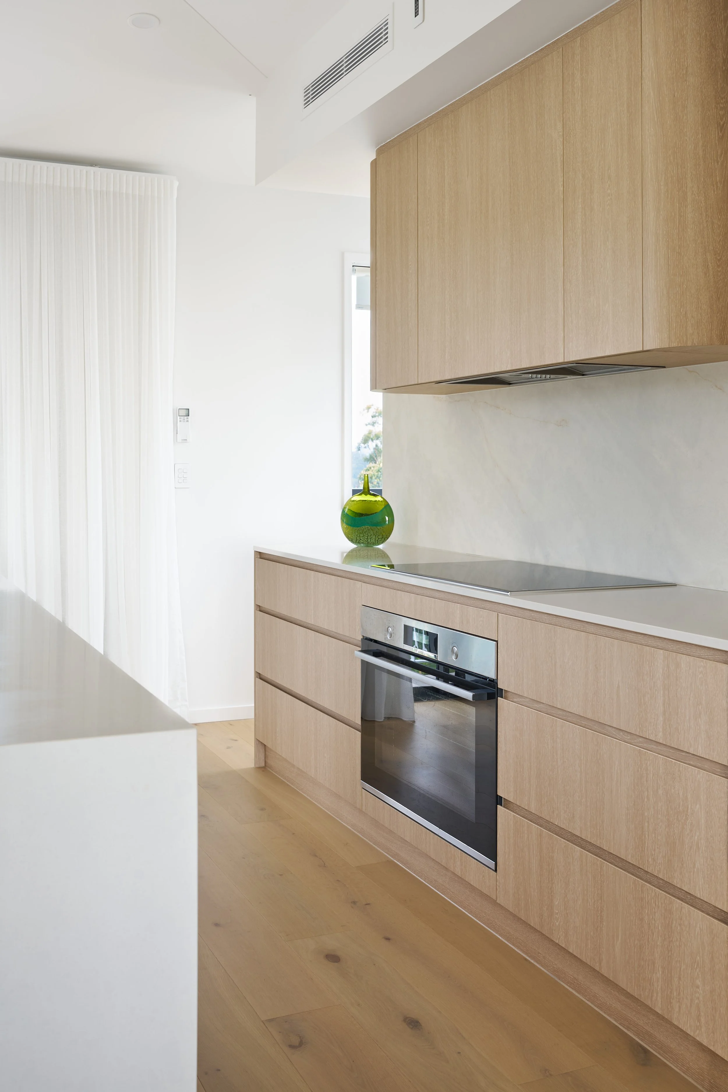Modern kitchen with light wooden cabinets, a built-in oven, and a white marble backsplash. A green decorative vase is placed on the counter, and there is natural light coming from a window.