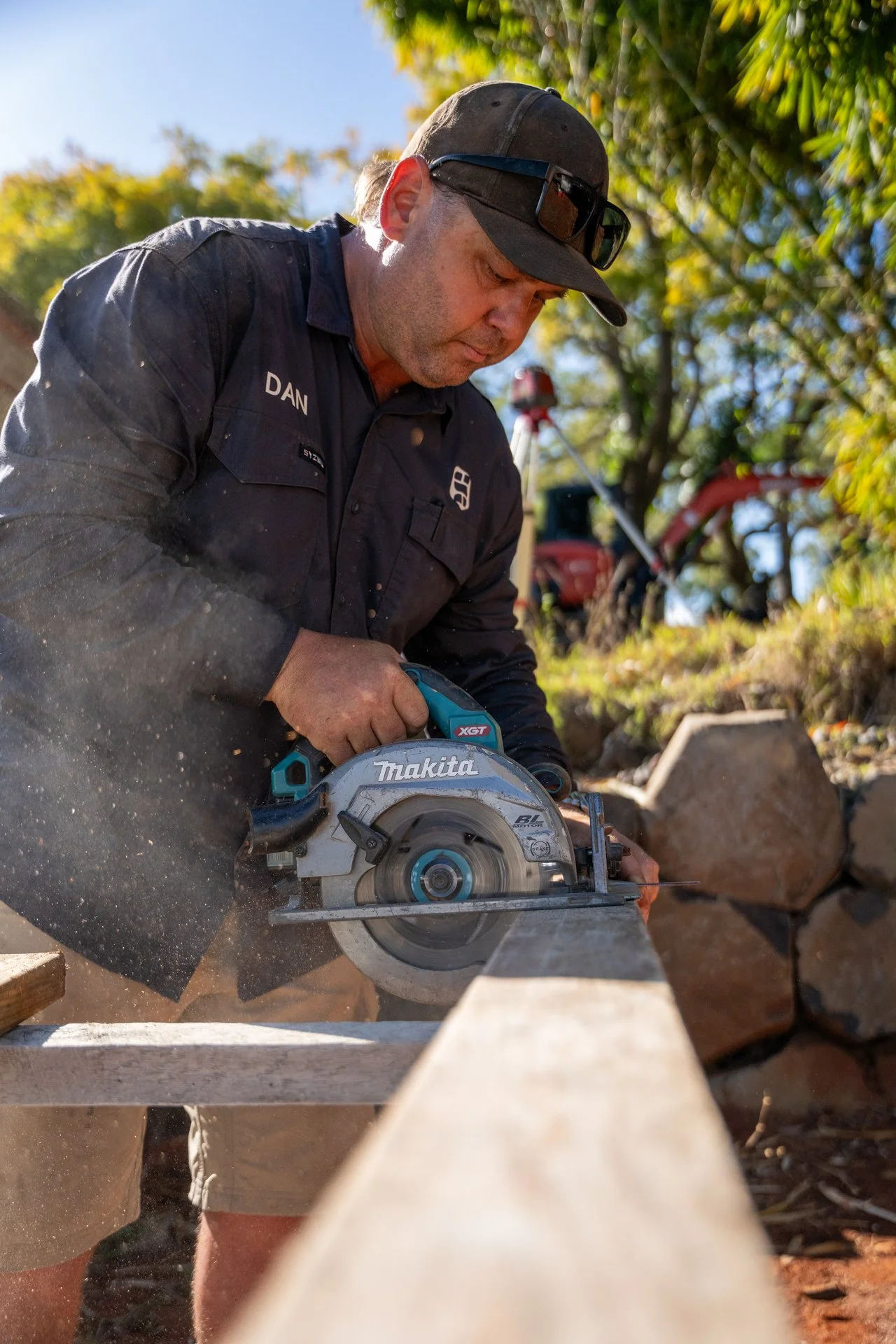 A man wearing sunglasses and a black shirt labeled 'DAN' is working outdoors using a circular saw to cut a piece of wood. The background has trees and machinery.