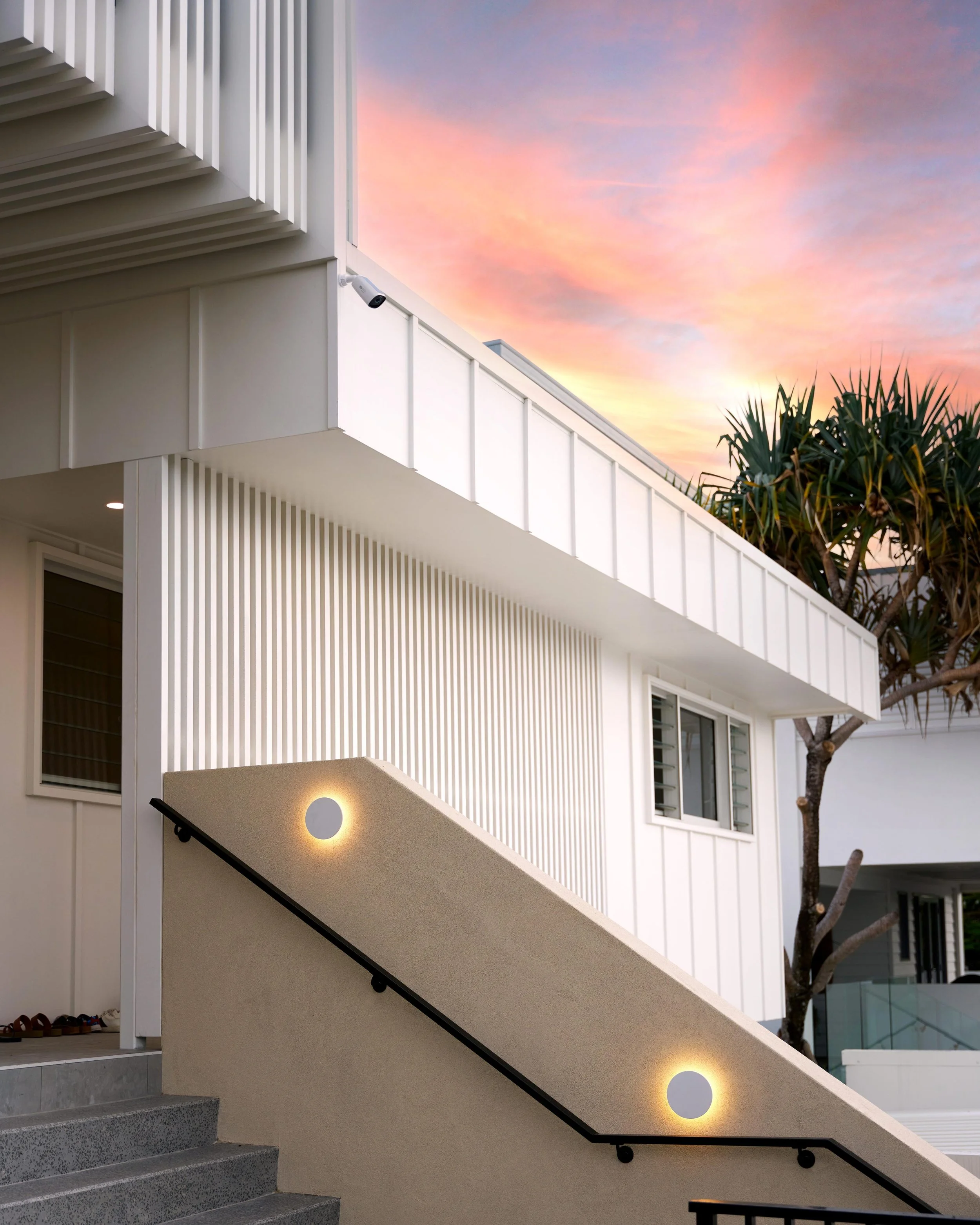 Exterior of a modern white building with vertical slats, three circular lights on the staircase wall, and a tree with spiky leaves against a colorful pink and purple sunset sky.