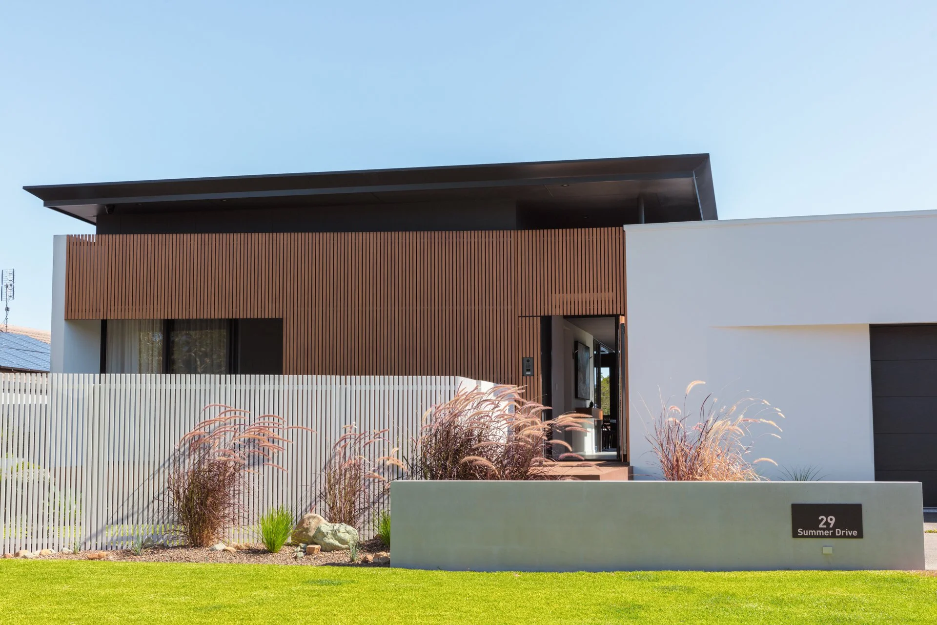 Modern house with a flat roof, wooden and white exterior walls, and a front yard with green grass and ornamental grasses, located at 29 Summer Drive.