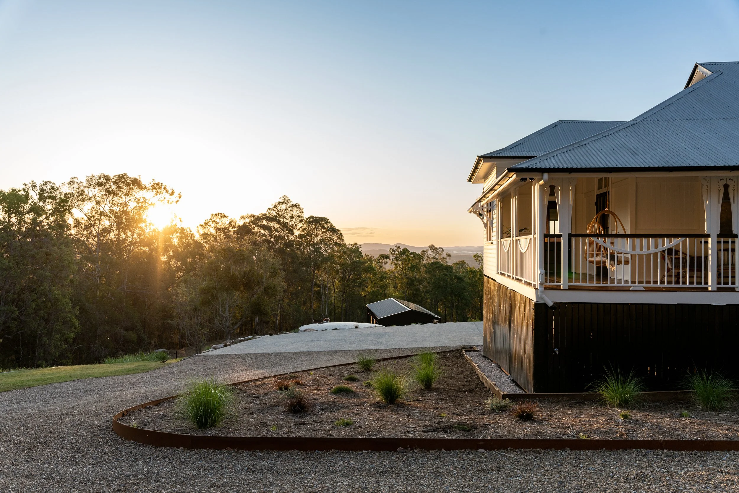 A house with a covered porch and railing, set in a landscaped yard with new grass and small plants, during sunset with trees and mountains in the background.