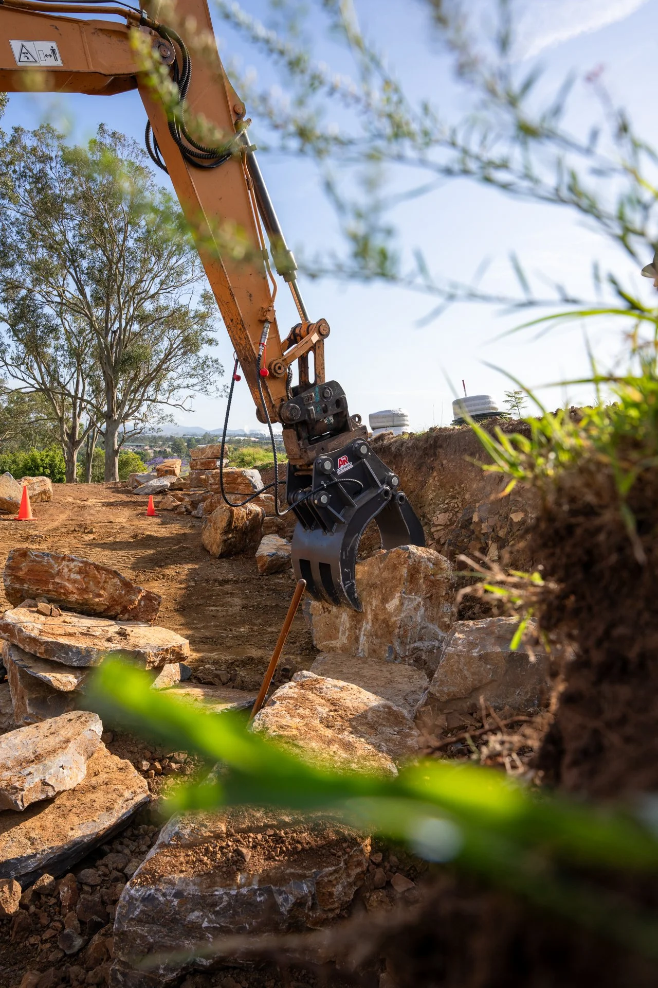 Close-up of a hydraulic excavator with a rock-breaking attachment working on a rocky area outdoors under a blue sky, with trees and orange traffic cones in the background.