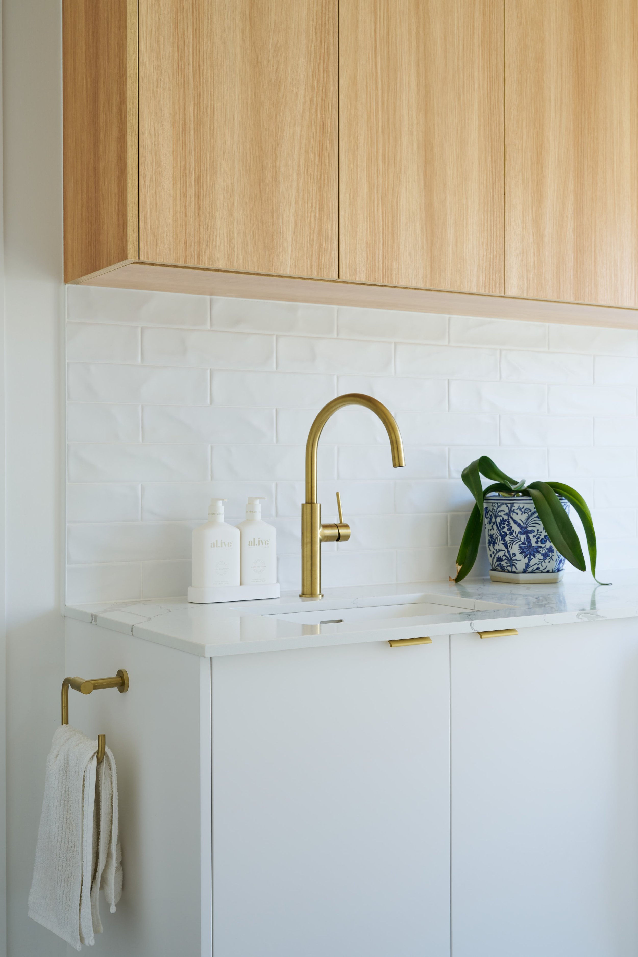A white bathroom vanity with a marble countertop and a gold faucet. Two white bottles labeled 'al.live' are on the counter. A potted plant with large, dark green leaves in a blue and white patterned pot is on the right. A towel hangs from a gold towe