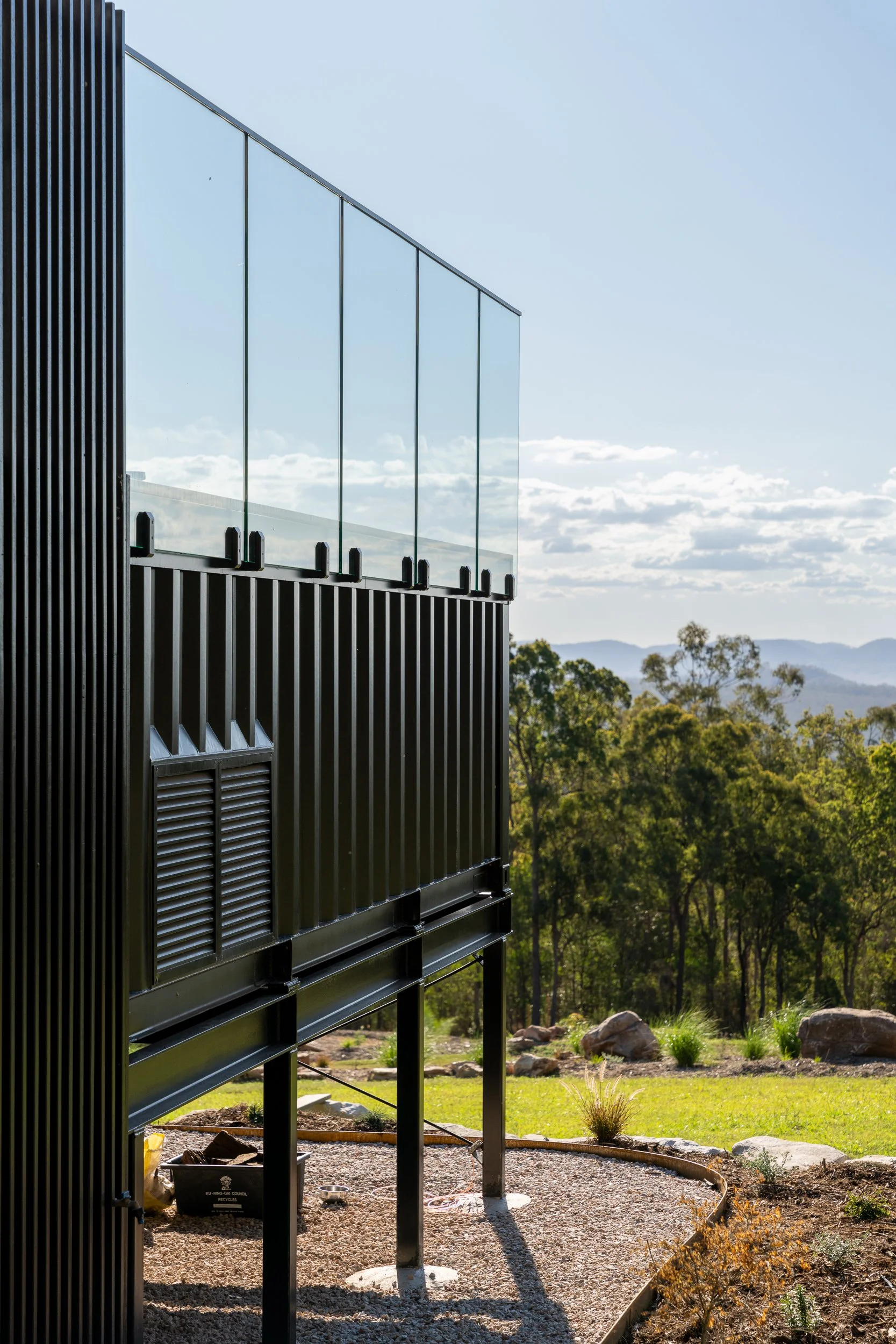 Modern building with black vertical panels and glass railing, overlooking a landscape with trees and mountains under a partly cloudy sky.