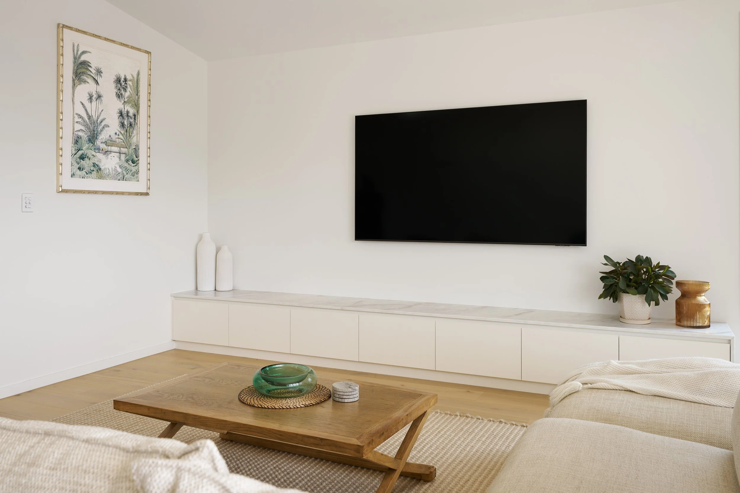 Living room with a wall-mounted flat screen TV, white sideboard, framed tropical artwork, two white vases, green plant in a beige basket, and a wooden coffee table with glass art and coasters.