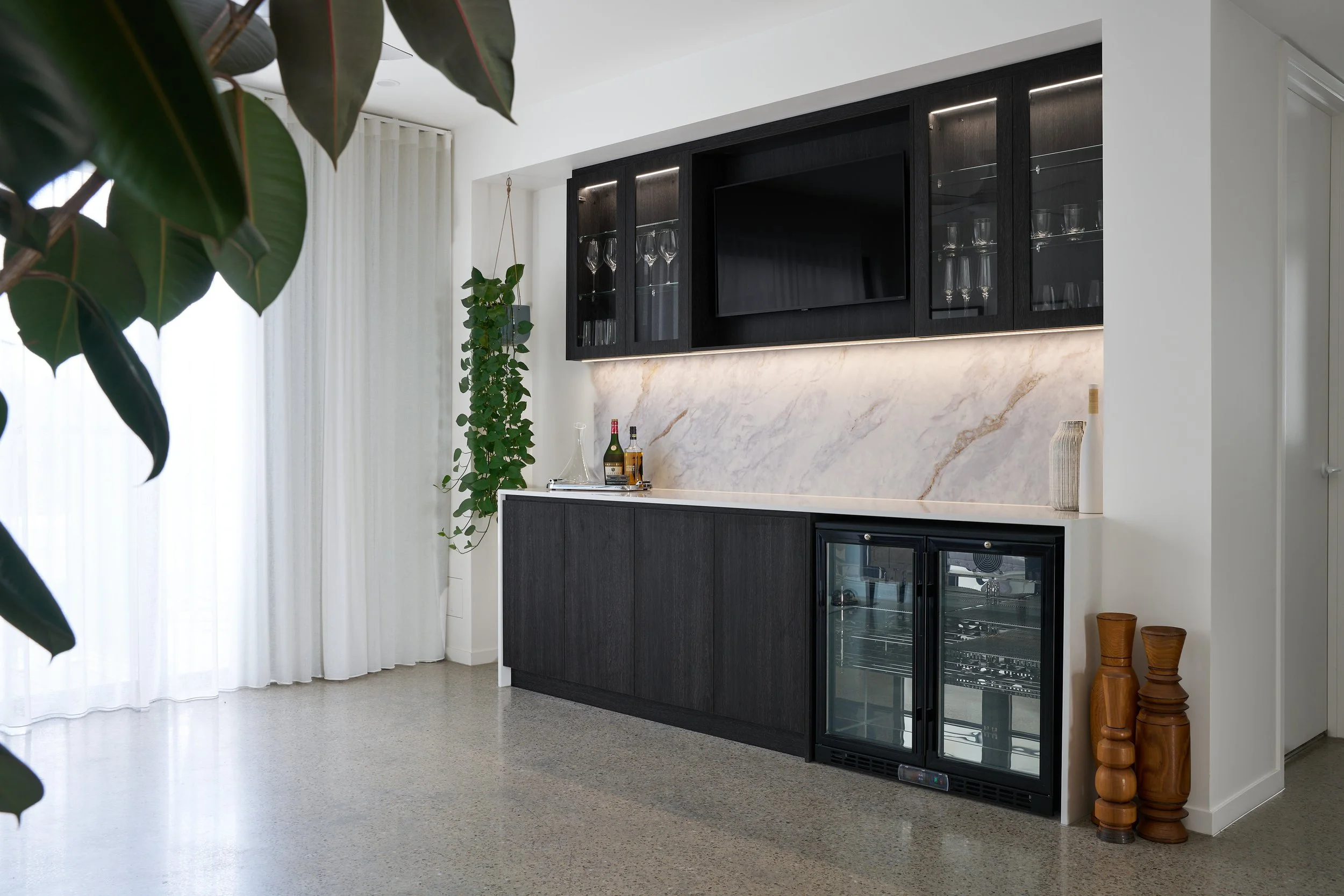A modern kitchen with black cabinets, a white marble backsplash, and a built-in TV. There are glassware inside the cabinets and a small bar area with bottles. A hanging potted plant is near a white curtain, and wooden decorative objects are on the fl