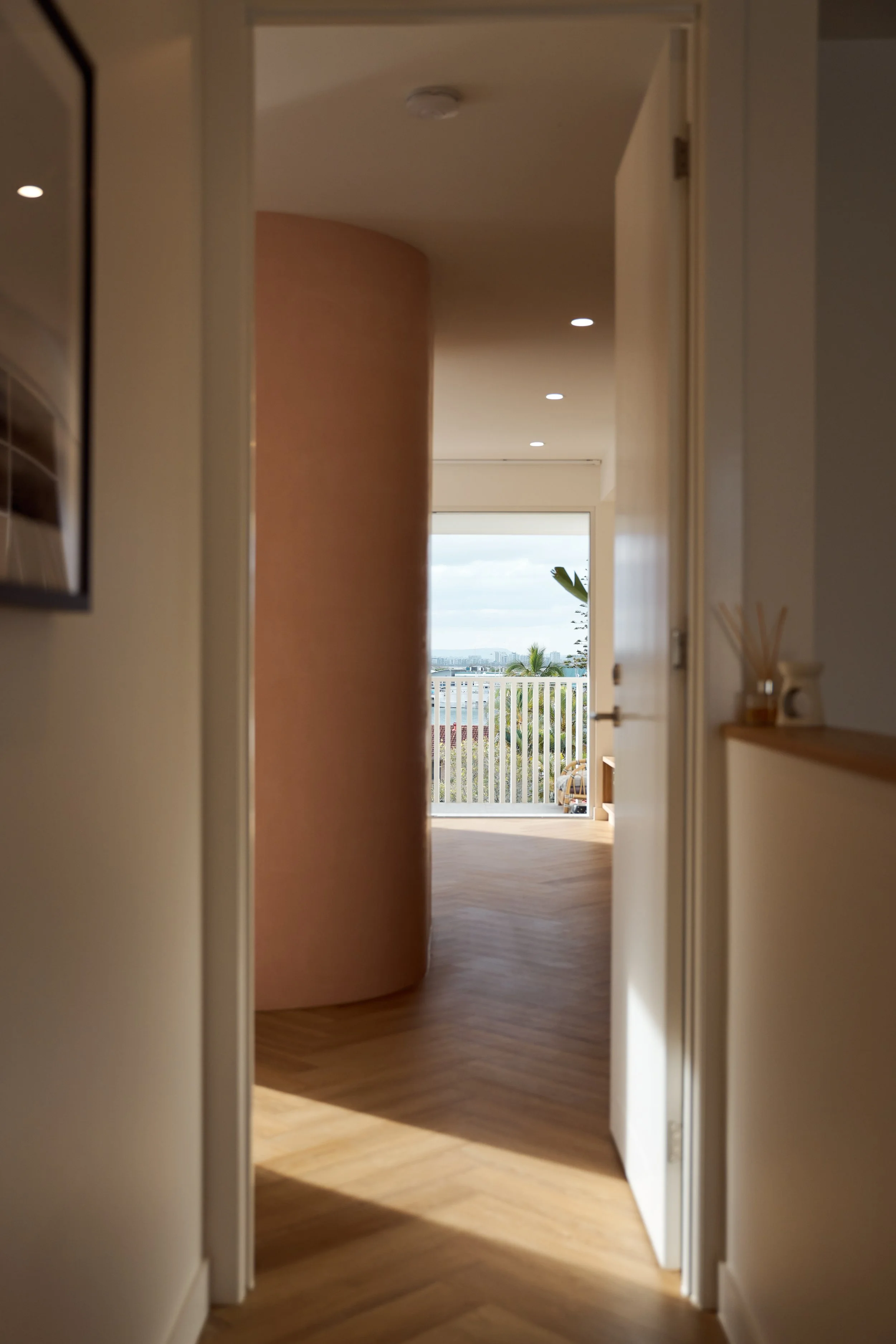 A hallway leading to a balcony with a view of the city and palm trees. The space has wooden flooring, white walls, and recessed lighting.