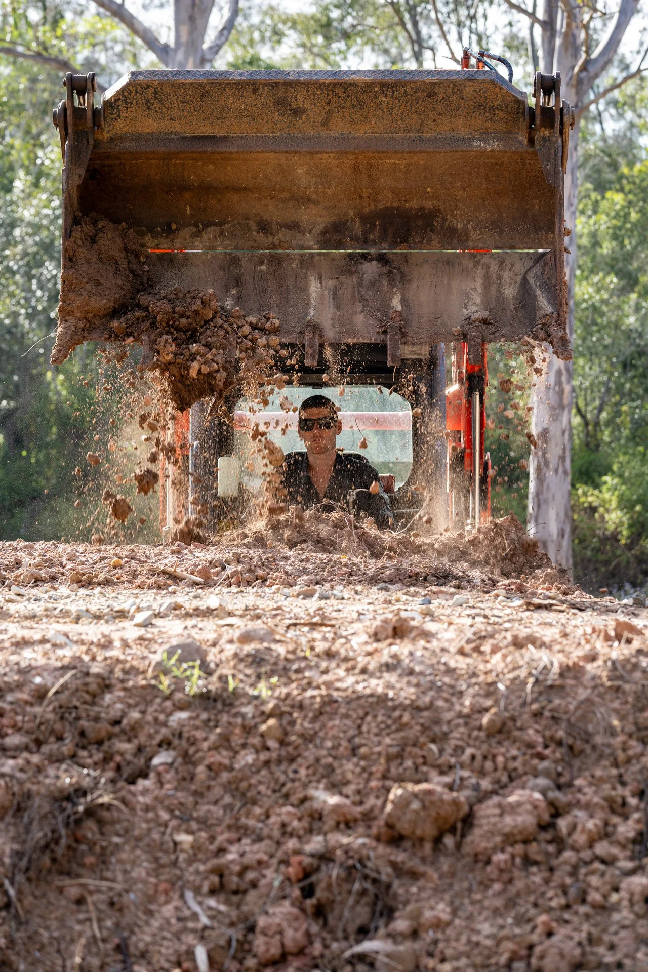 A person operating a bulldozer on a dirt and gravel surface, with trees and sunlight in the background.